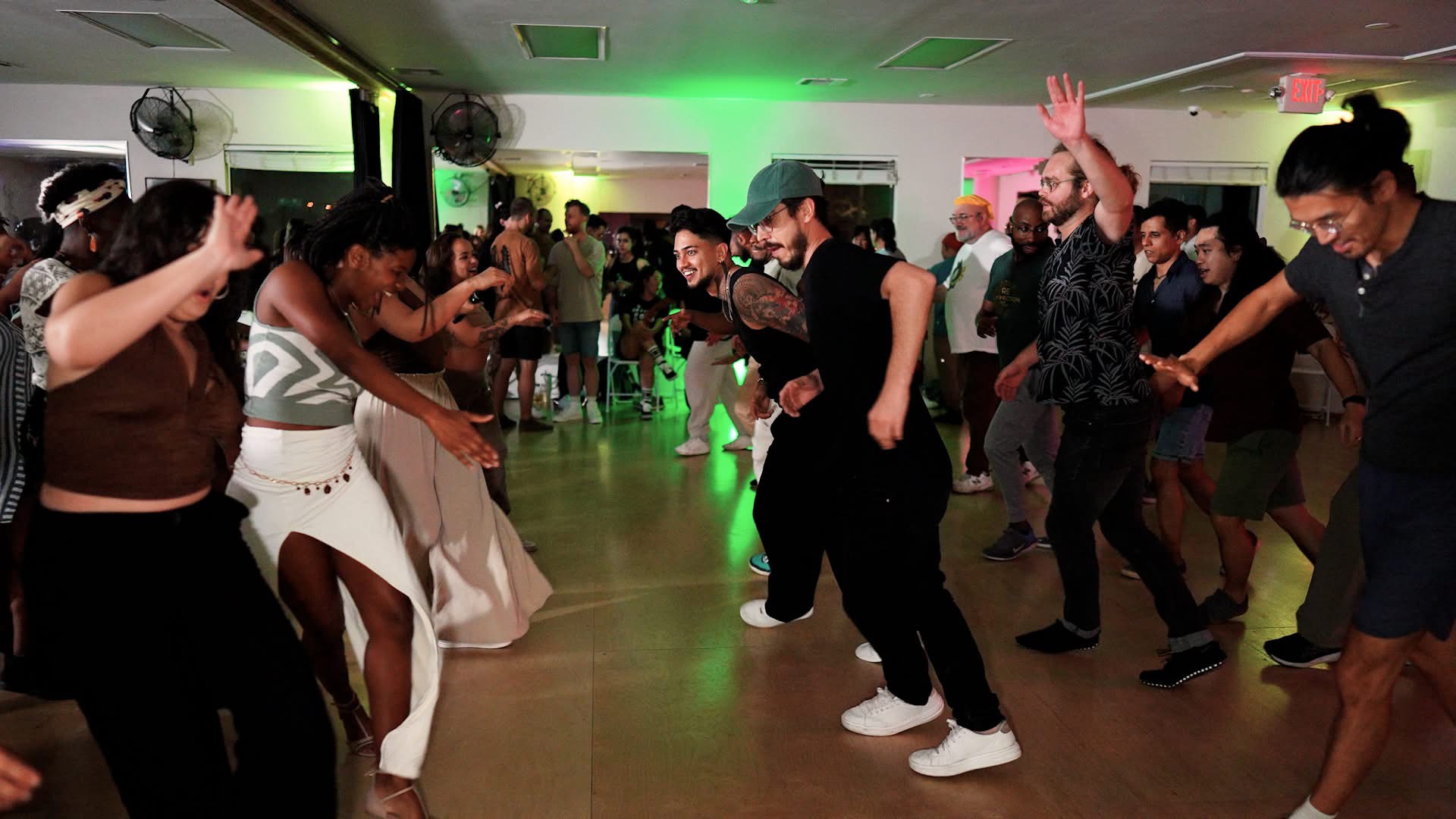 People dancing in a room with green and pink lighting, some with hands raised, enjoying a social event.