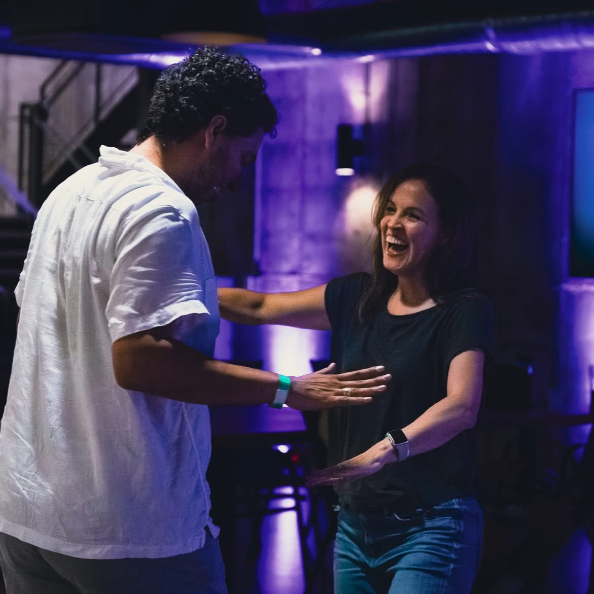 Two people smiling and interacting in a dimly lit room with purple lighting, one with curly hair and the other with straight hair.