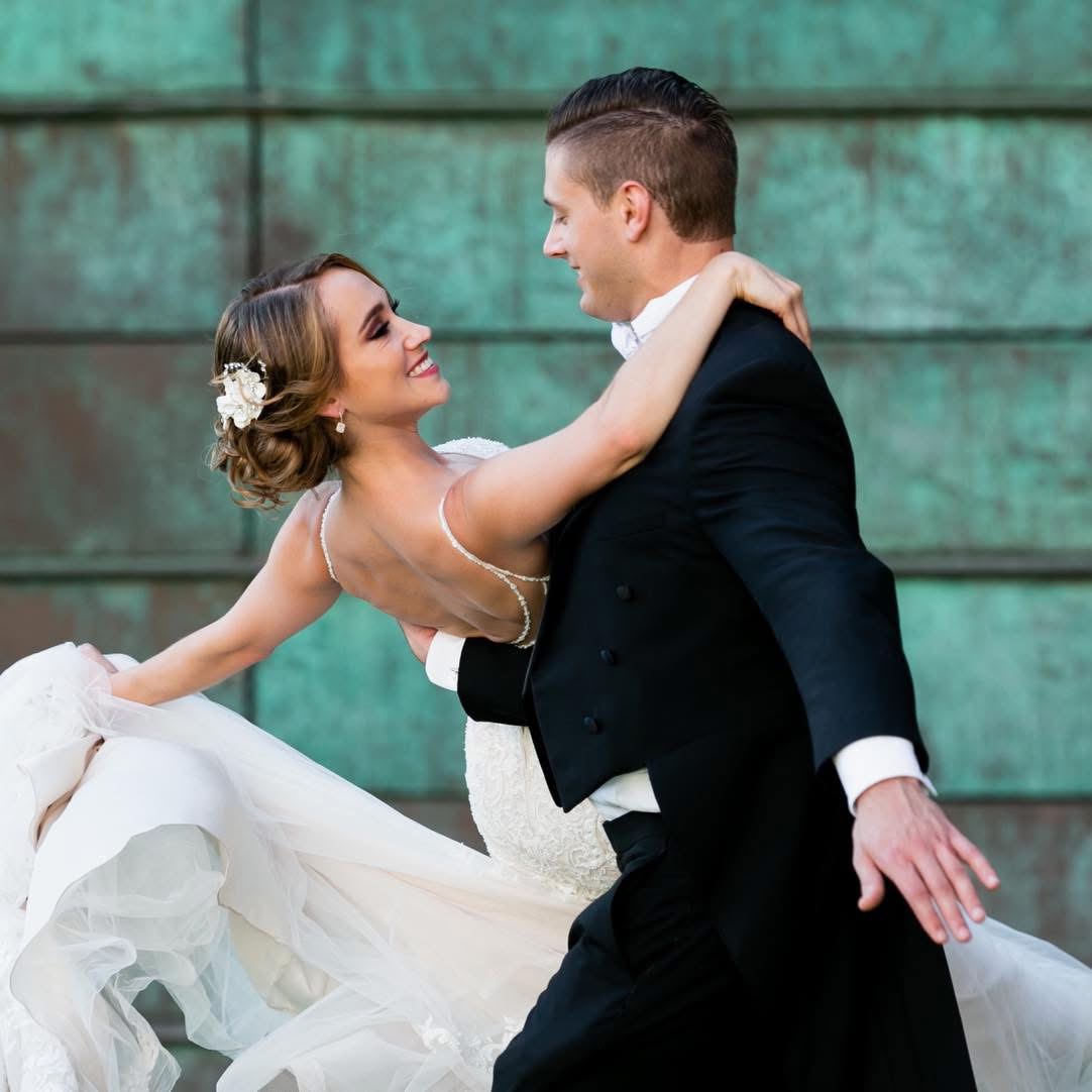 A bride and groom dance outdoors, smiling at each other, with the bride in a white gown and the groom in a black suit.