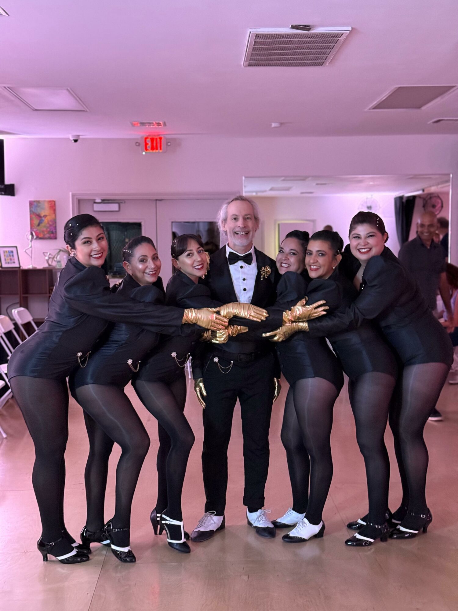 Group of six women in black outfits and a man in a tuxedo, all holding gold gloves, posing indoors.