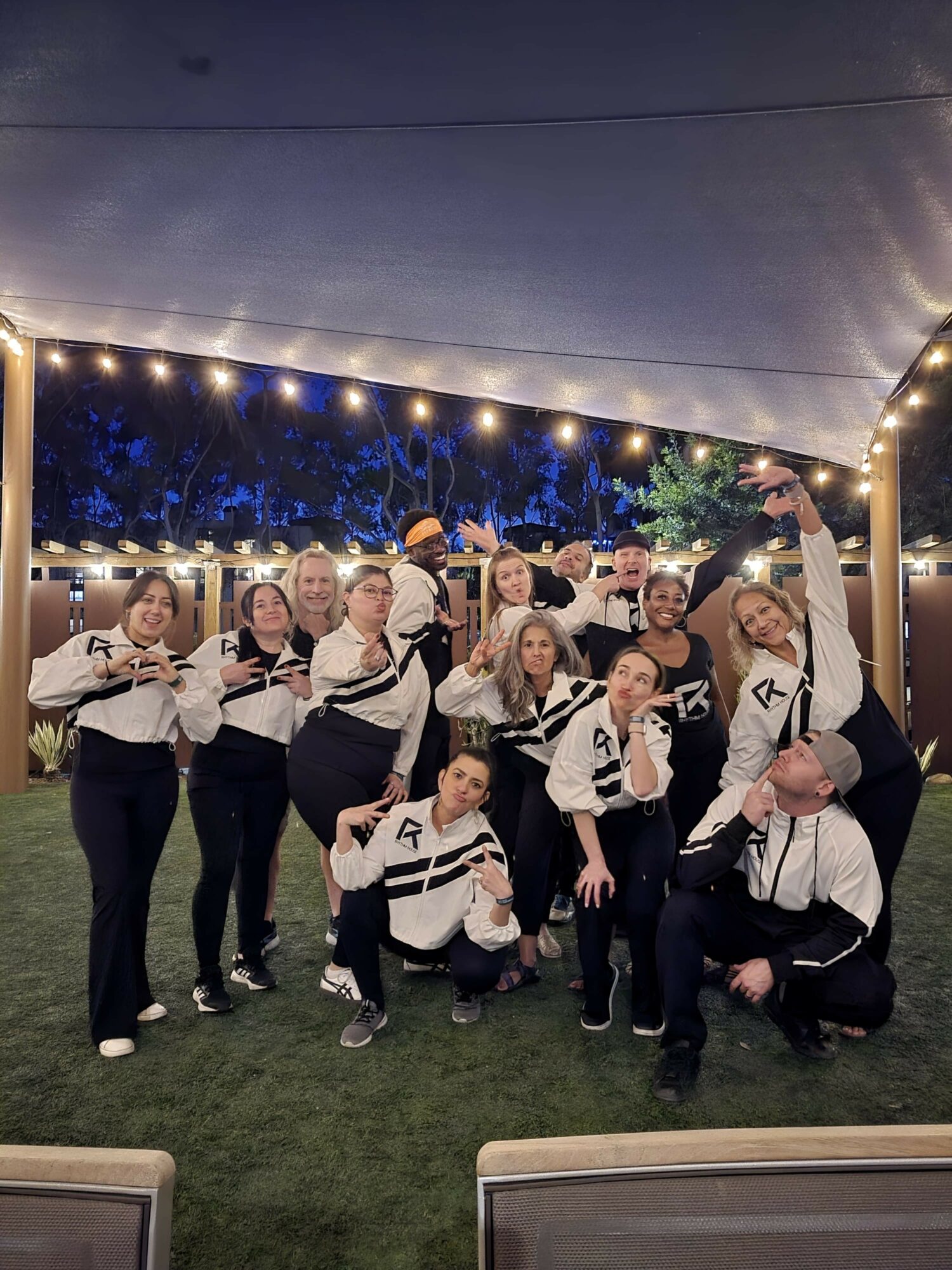 Group of people posing outdoors under string lights at night, some making hand gestures, with trees in the background.