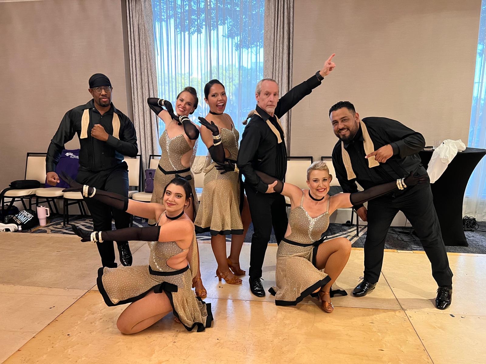 Group of seven dancers in costumes posing indoors with a beige wall and window behind them.