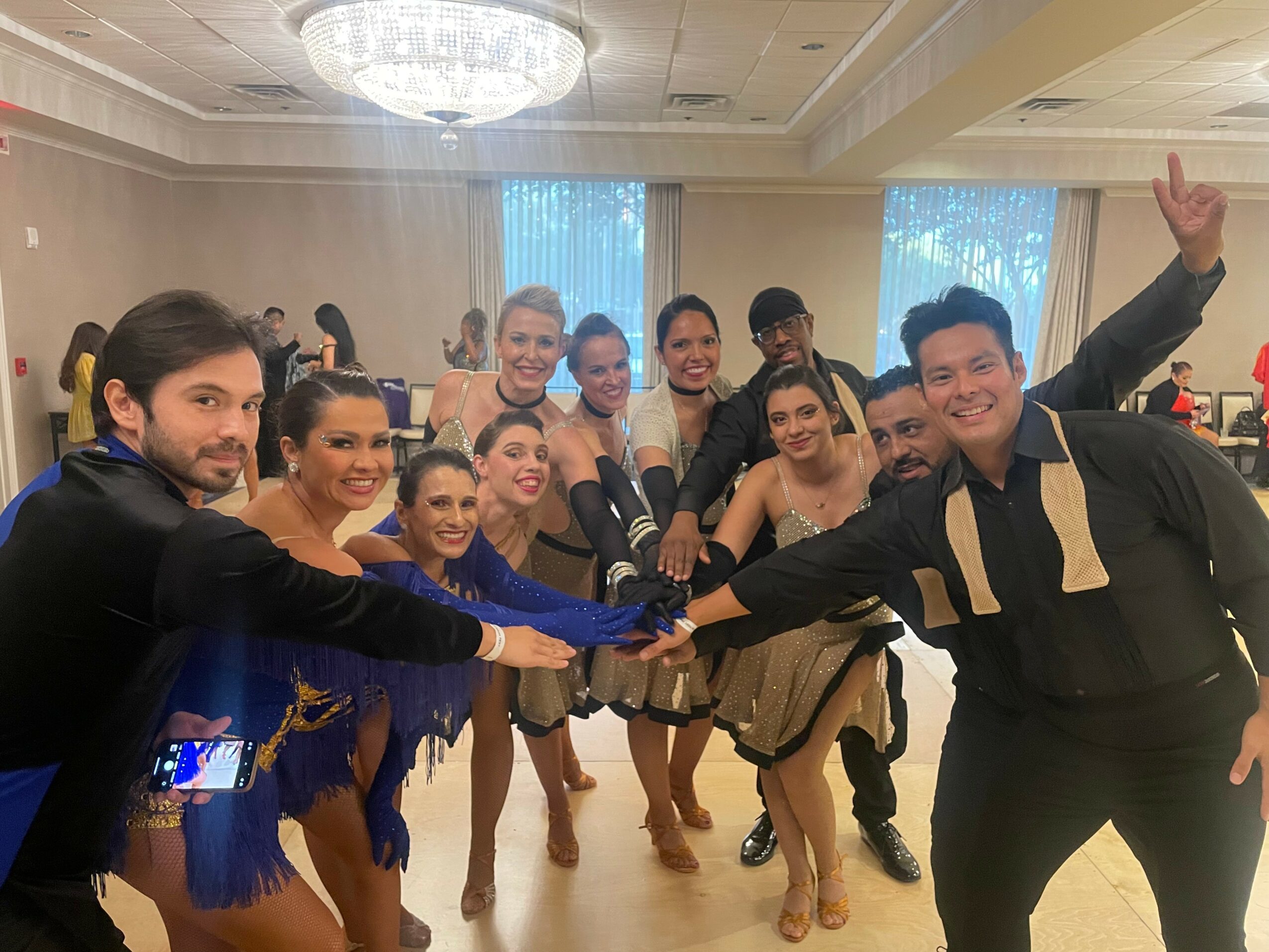 Group of people in dance costumes smiling and posing in a decorated room, some with hands joined in the center.