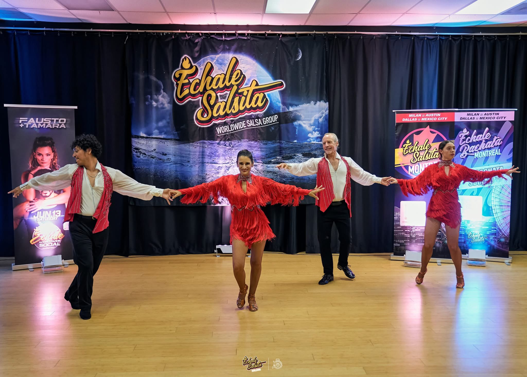 Four dancers perform on stage with a backdrop and banners, holding hands and wearing red and white costumes.