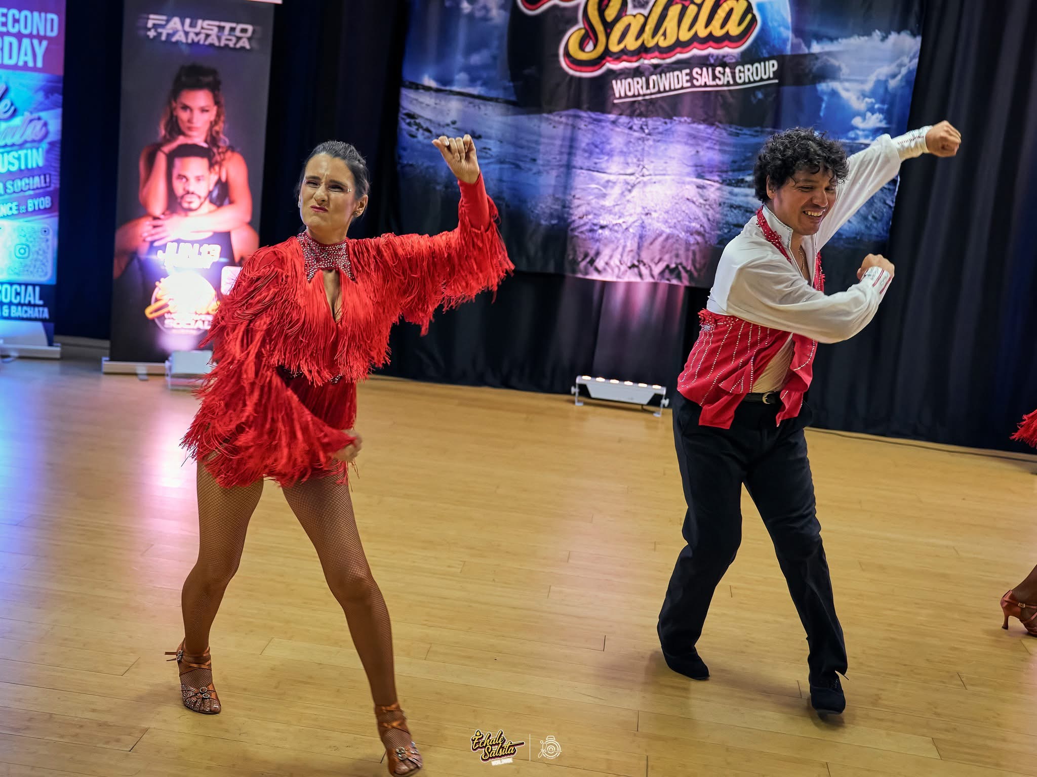 Two dancers perform on stage, one woman in a red feathered costume and one man in a red and white outfit, with a backdrop and posters behind them.