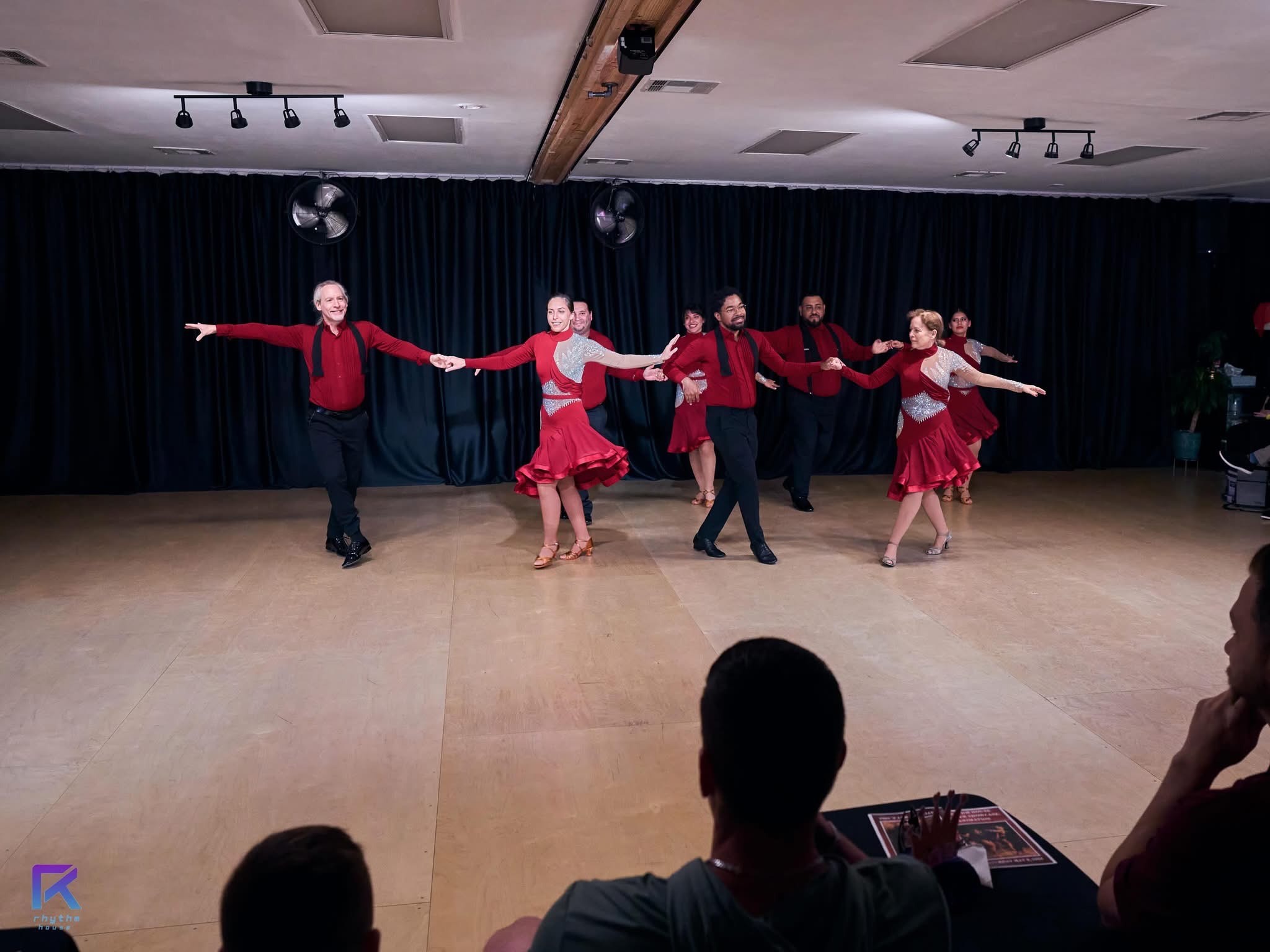 Five dancers in red costumes perform on a stage with a dark curtain background, arms extended outward, in a dance pose.
