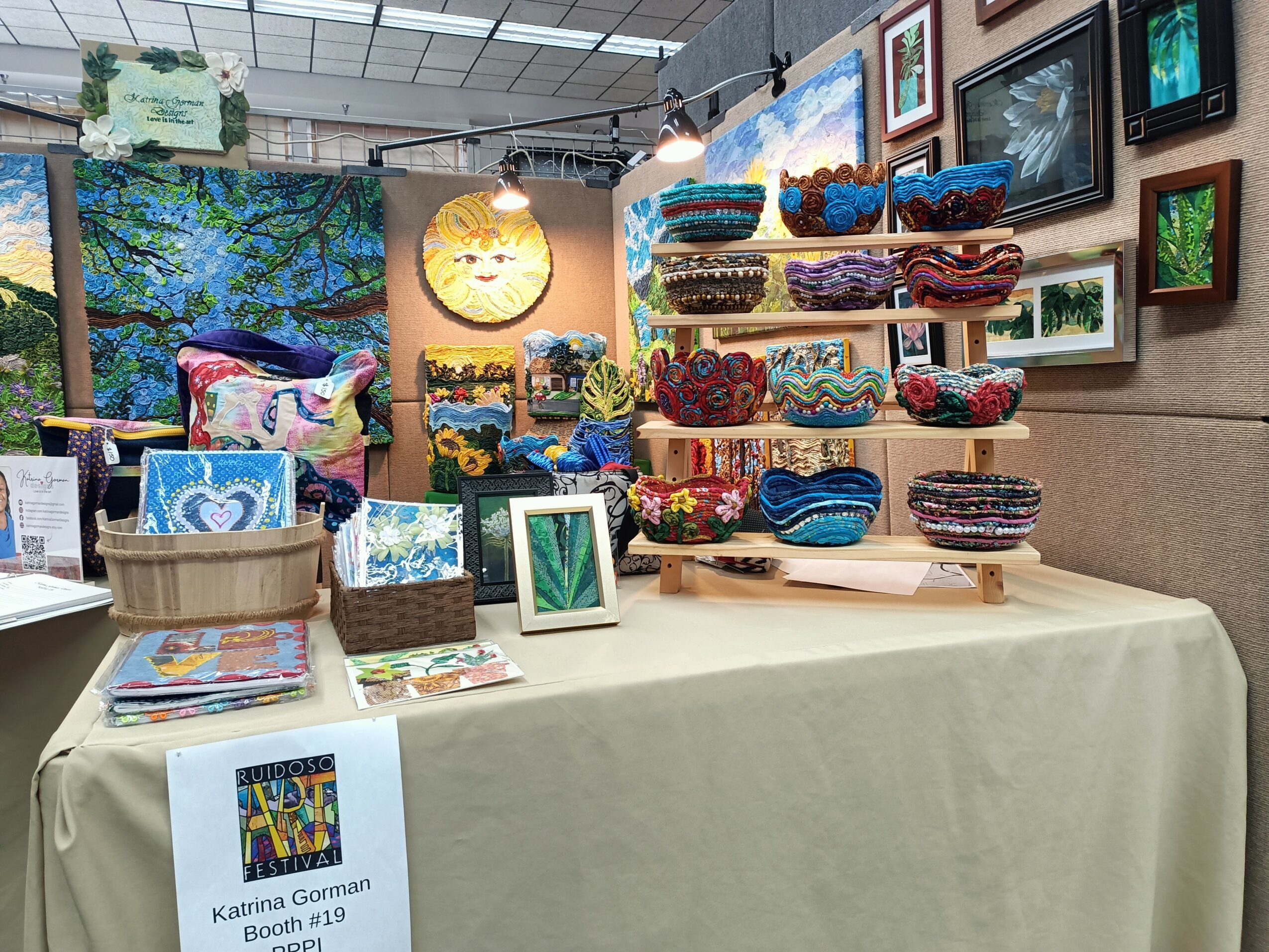 Display of colorful handmade textiles and framed artwork at an indoor craft booth, with a table covered in a beige cloth.