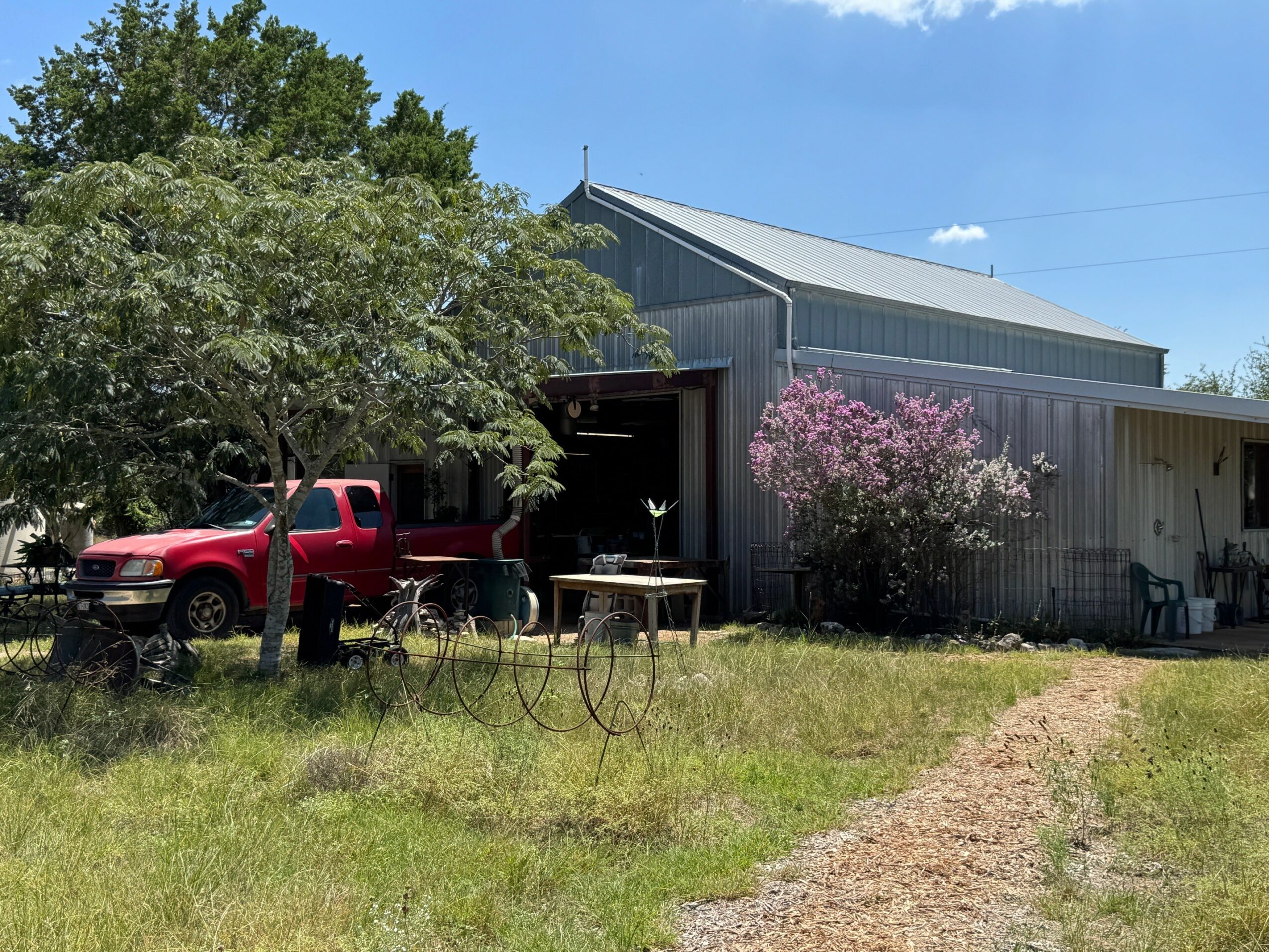 A metal barn with a tree and pink flowering bush in front, a red truck parked nearby, and outdoor furniture on grass.