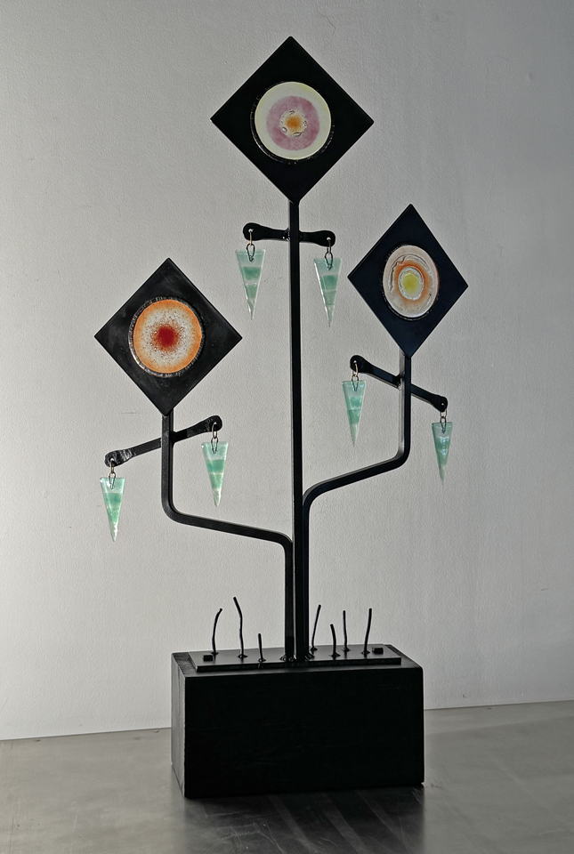 Sculpture with three diamond-shaped frames on a black stand, each containing colorful circular designs, with small green and white objects hanging below.