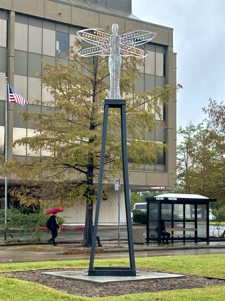 Large dragonfly sculpture on a tall black stand in a park with trees and a building in the background.