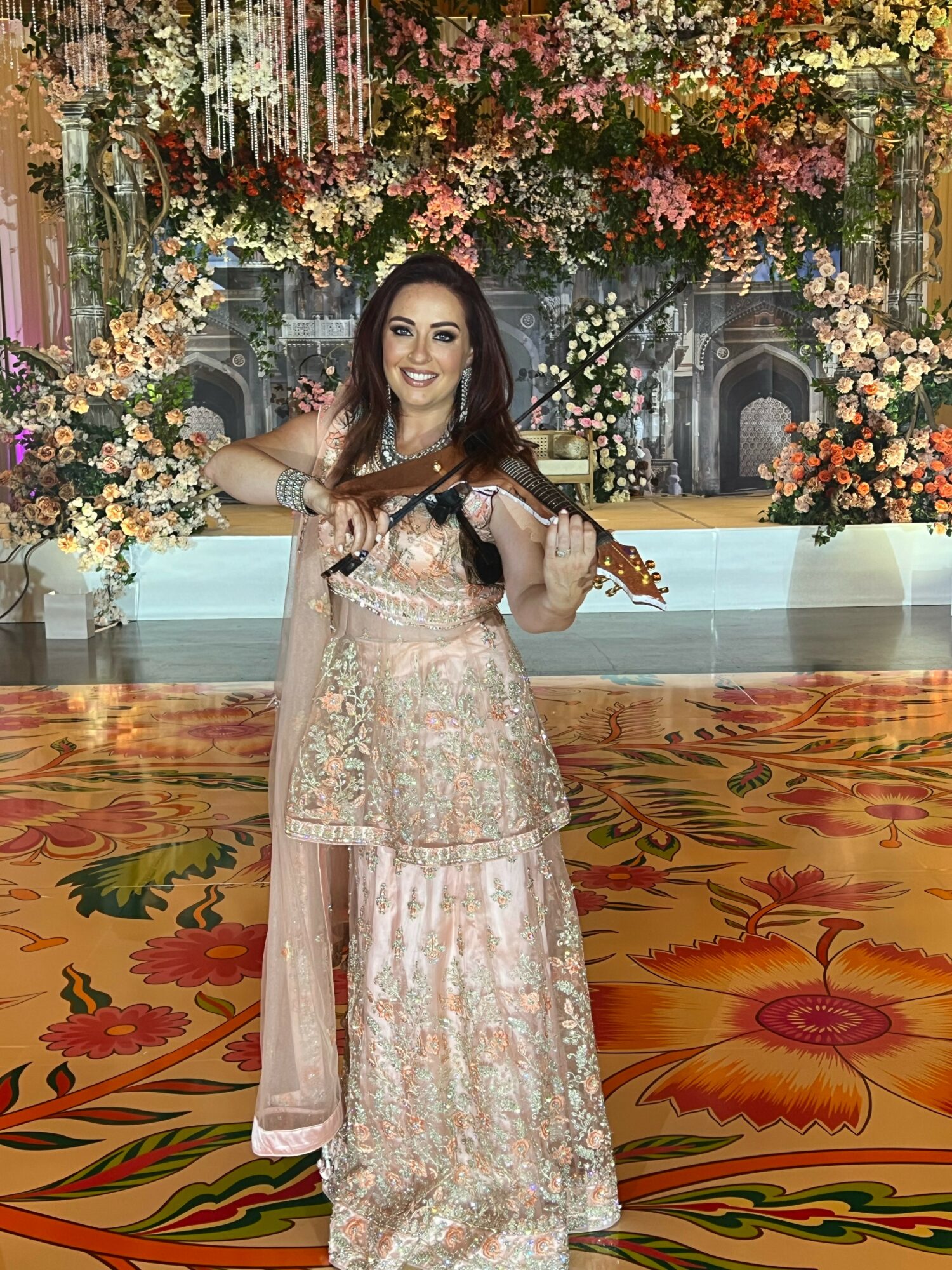 Woman in traditional attire holding a violin, standing in front of floral decorations and ornate background, smiling.