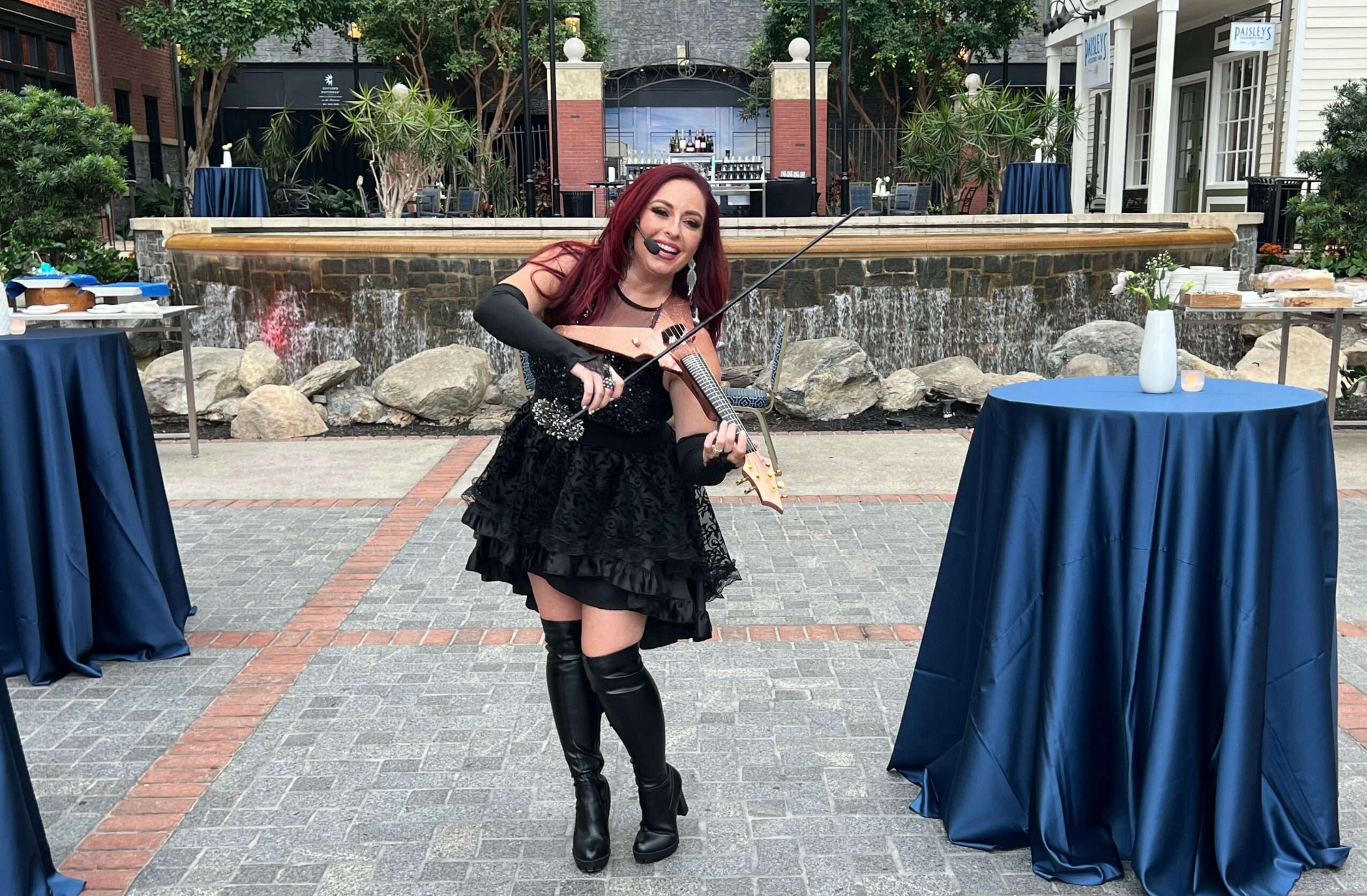 Young woman with red hair playing violin outdoors, standing between two tall tables with blue tablecloths, in front of a fountain.