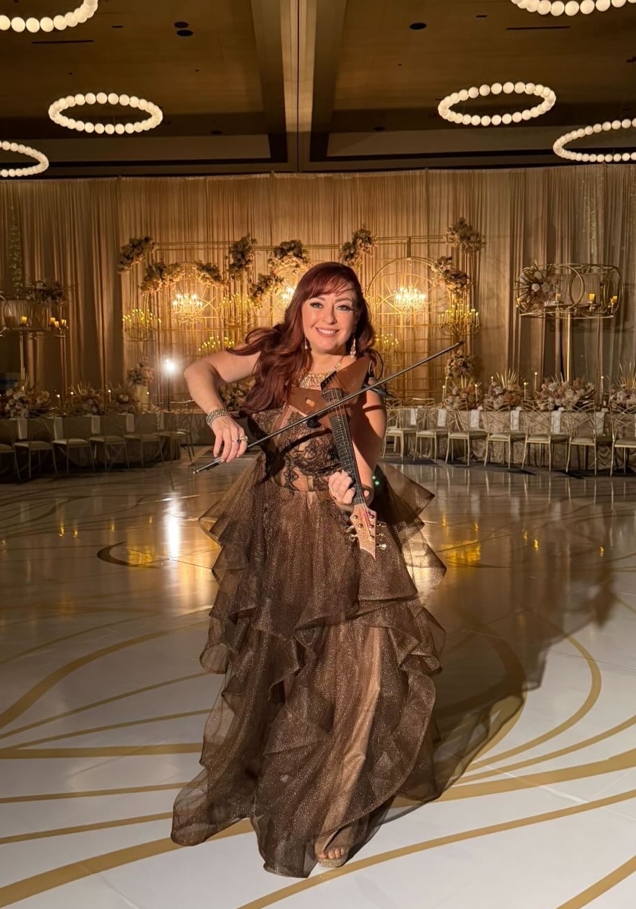 Woman in a flowing gown holding a violin, standing on a decorated dance floor with elegant lighting and backdrop.