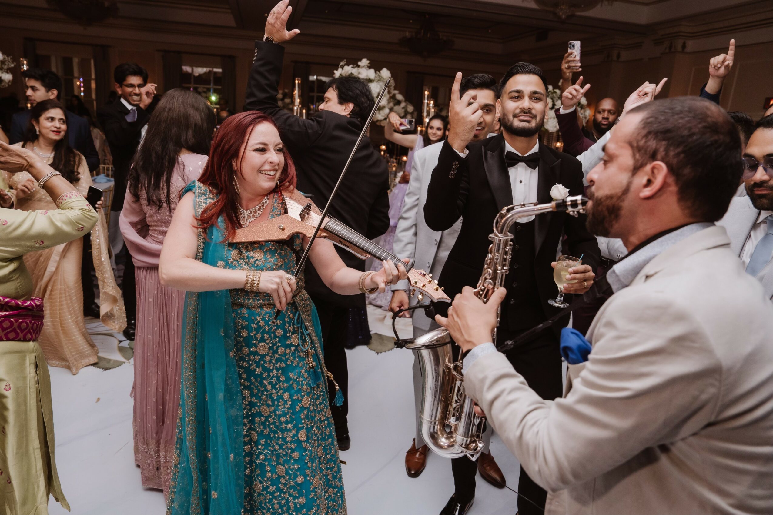 People at a celebration, woman playing violin, man playing saxophone, others dancing and smiling, festive atmosphere, formal attire.