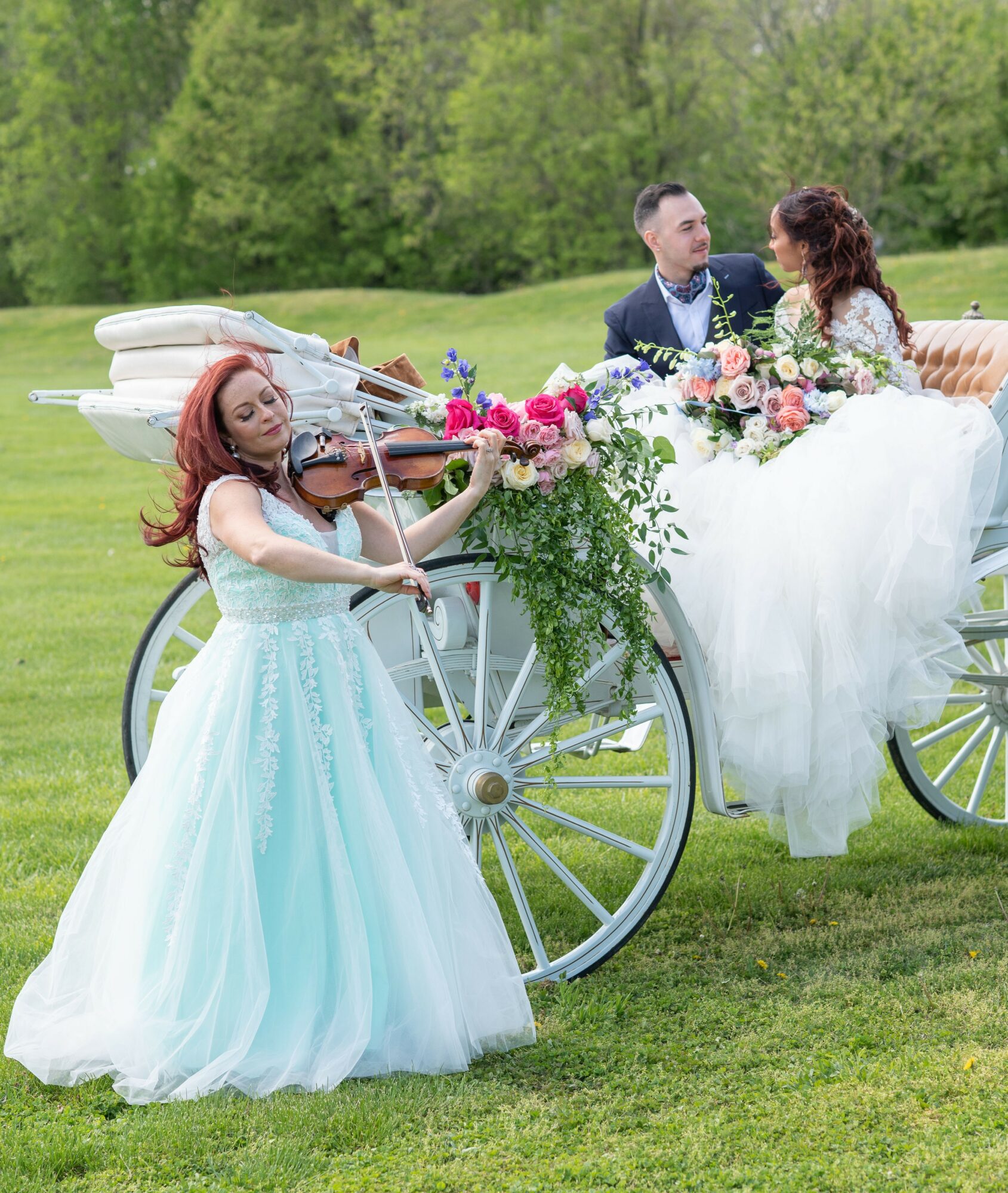 A woman in a light blue gown plays violin next to a decorated horse-drawn carriage with a bride and groom inside, outdoors.