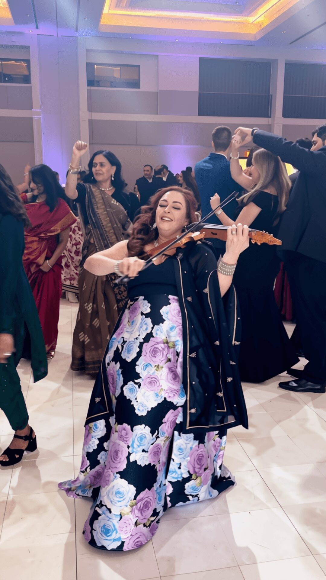 Woman playing violin, smiling, in a floral dress and black jacket, at a social event with people dancing in background.