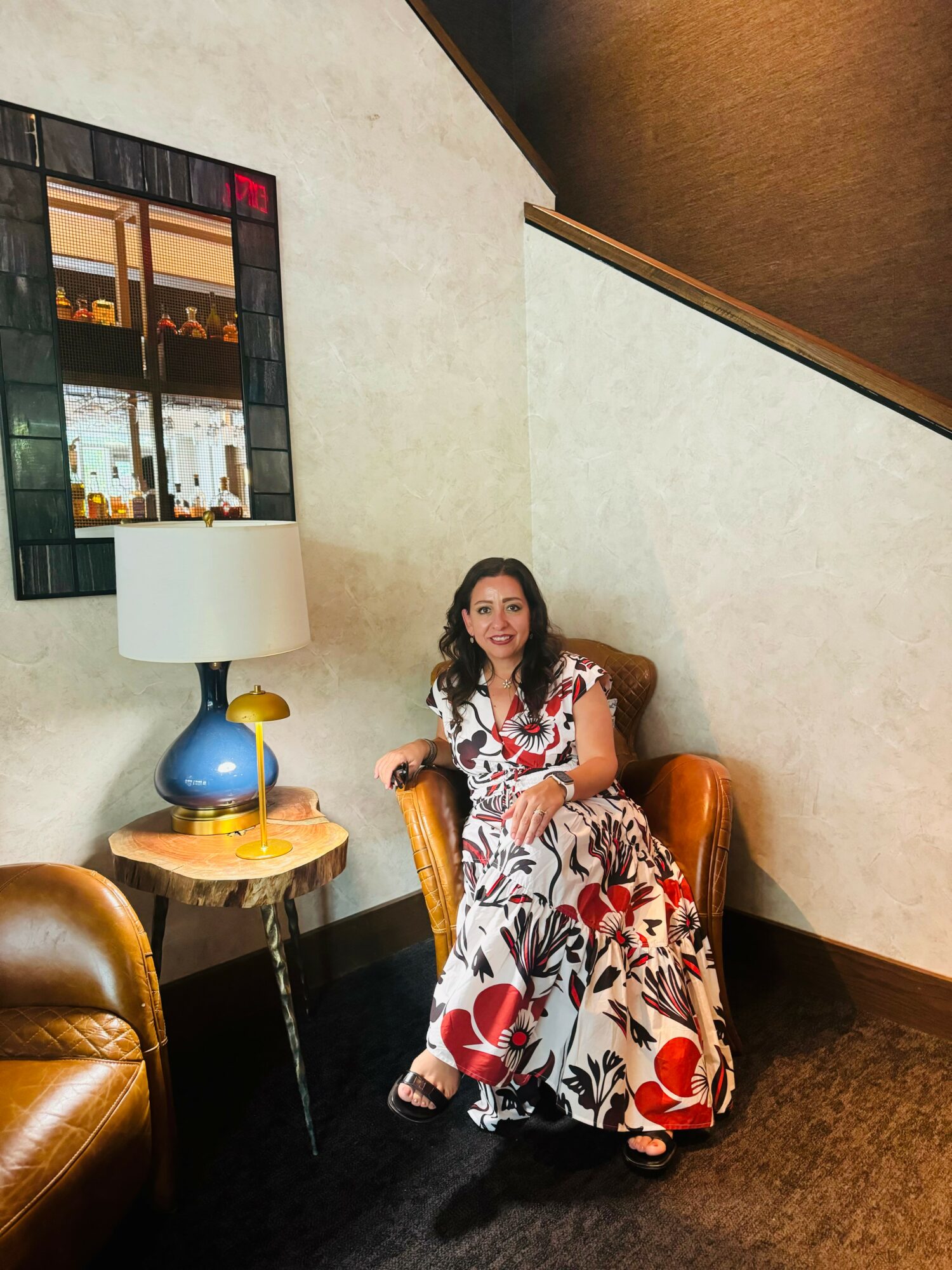 Woman in a floral dress sitting in a leather armchair in a room with a side table, lamp, and mirror.