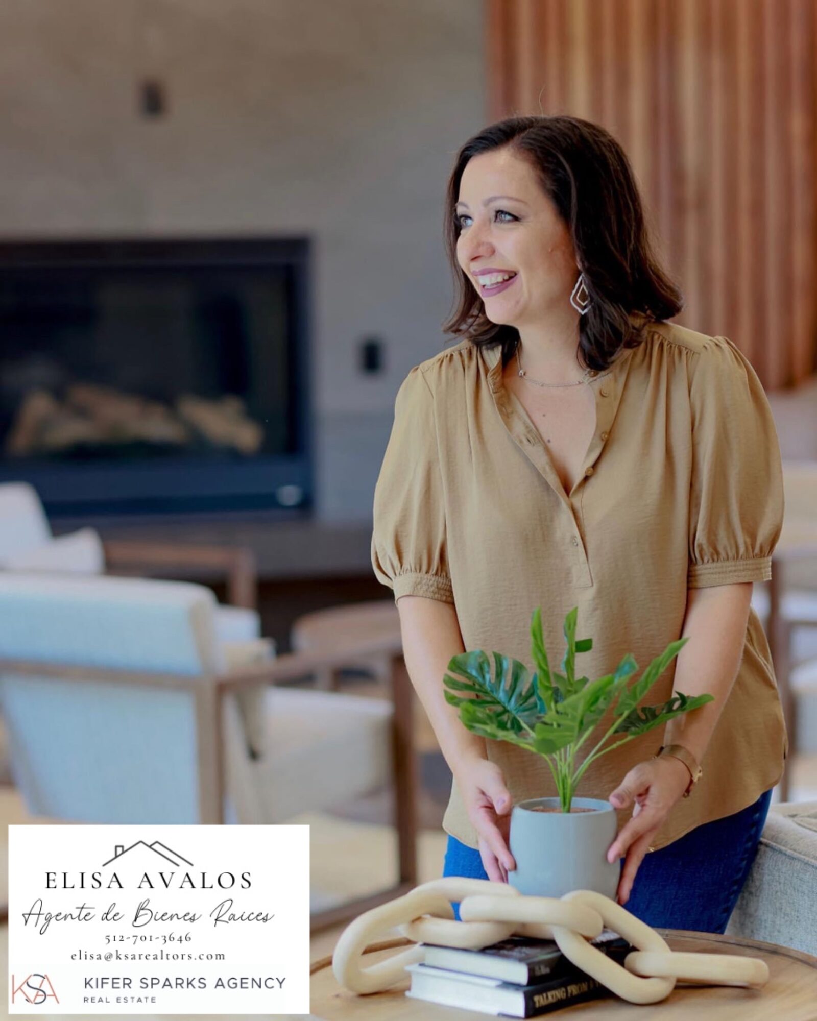 Woman smiling, holding a potted plant, standing in a living room with a fireplace and furniture.