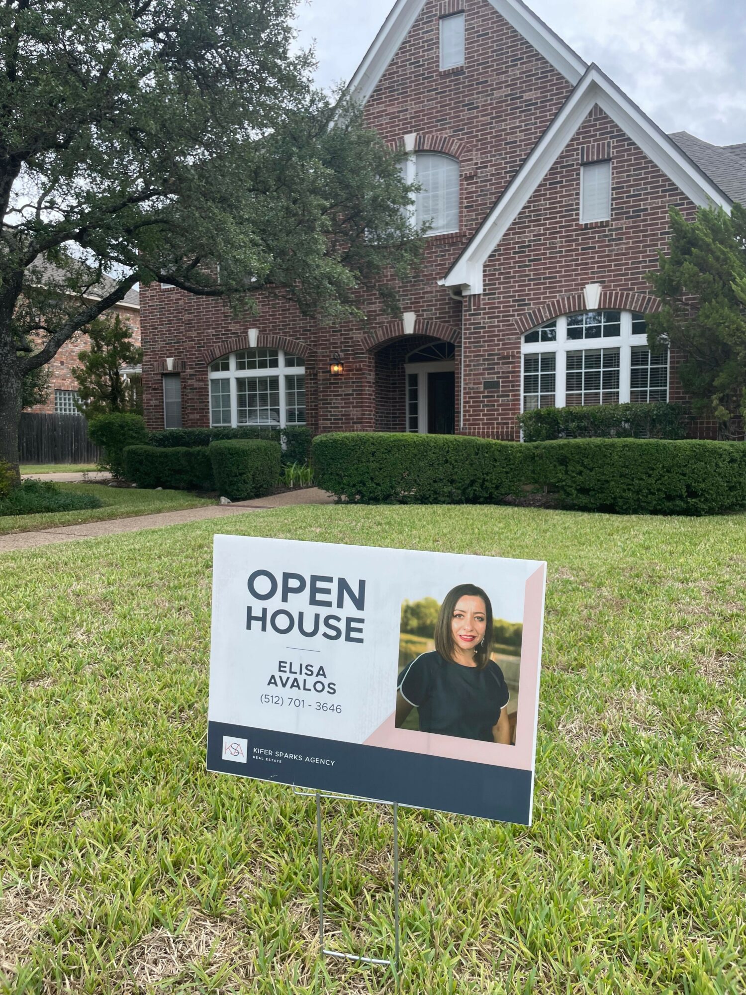 A sign in front of a brick house with white windows and a garden, indicating an open house event.