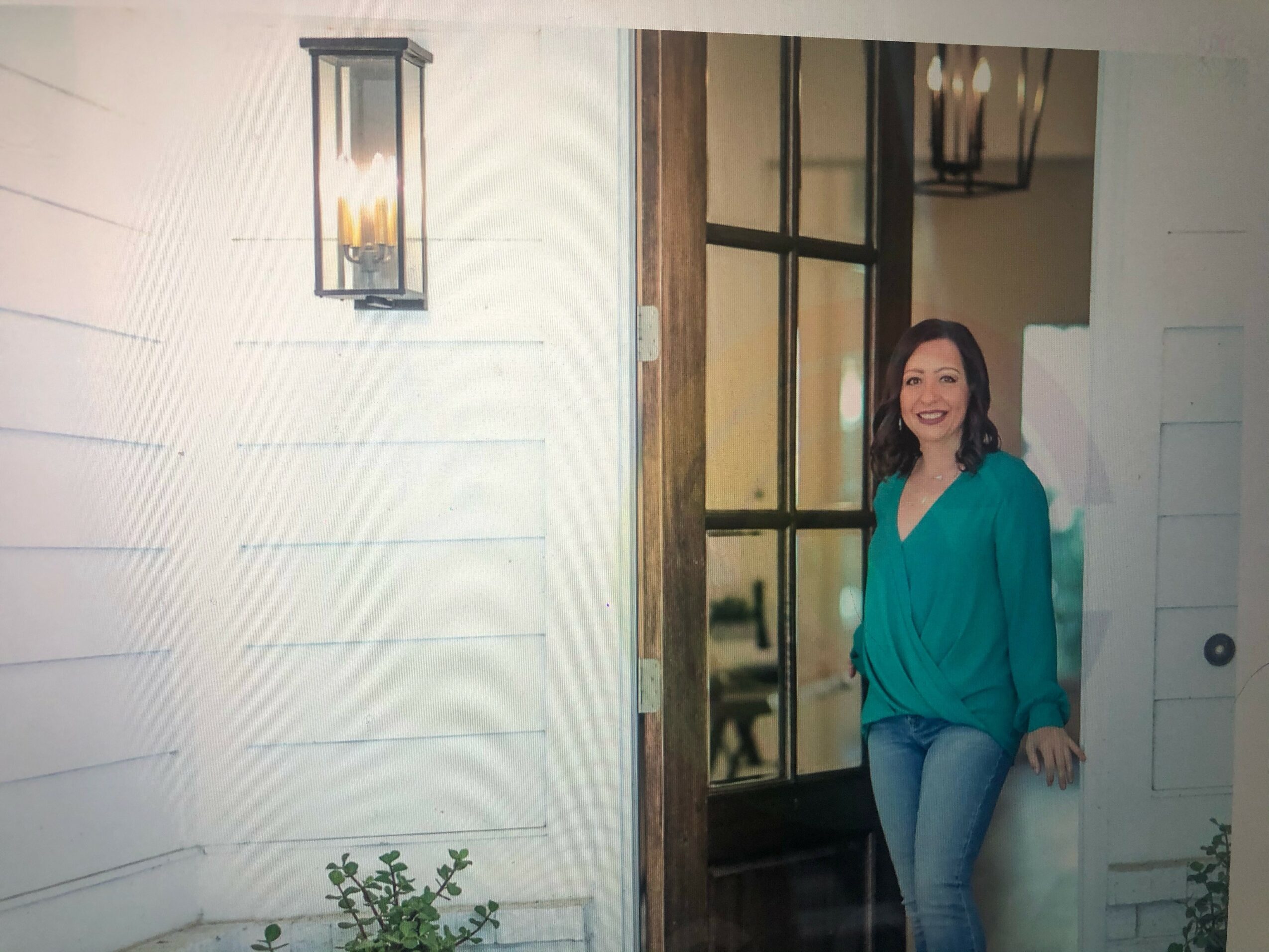 Woman with dark hair in a teal top and jeans standing outside near a door, smiling, with a wall-mounted light nearby.