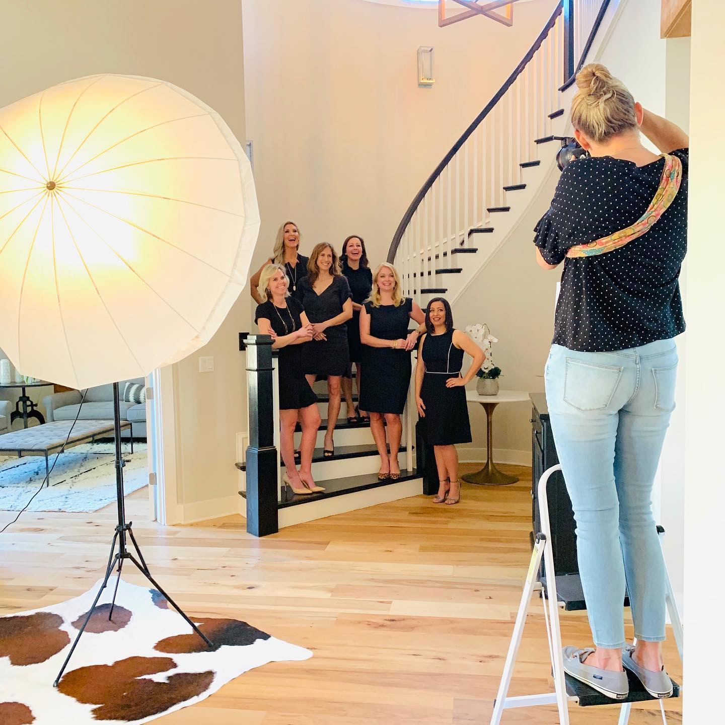 A woman takes a photo of six women standing on stairs in a home, with a large umbrella light nearby, hardwood floor, and staircase.