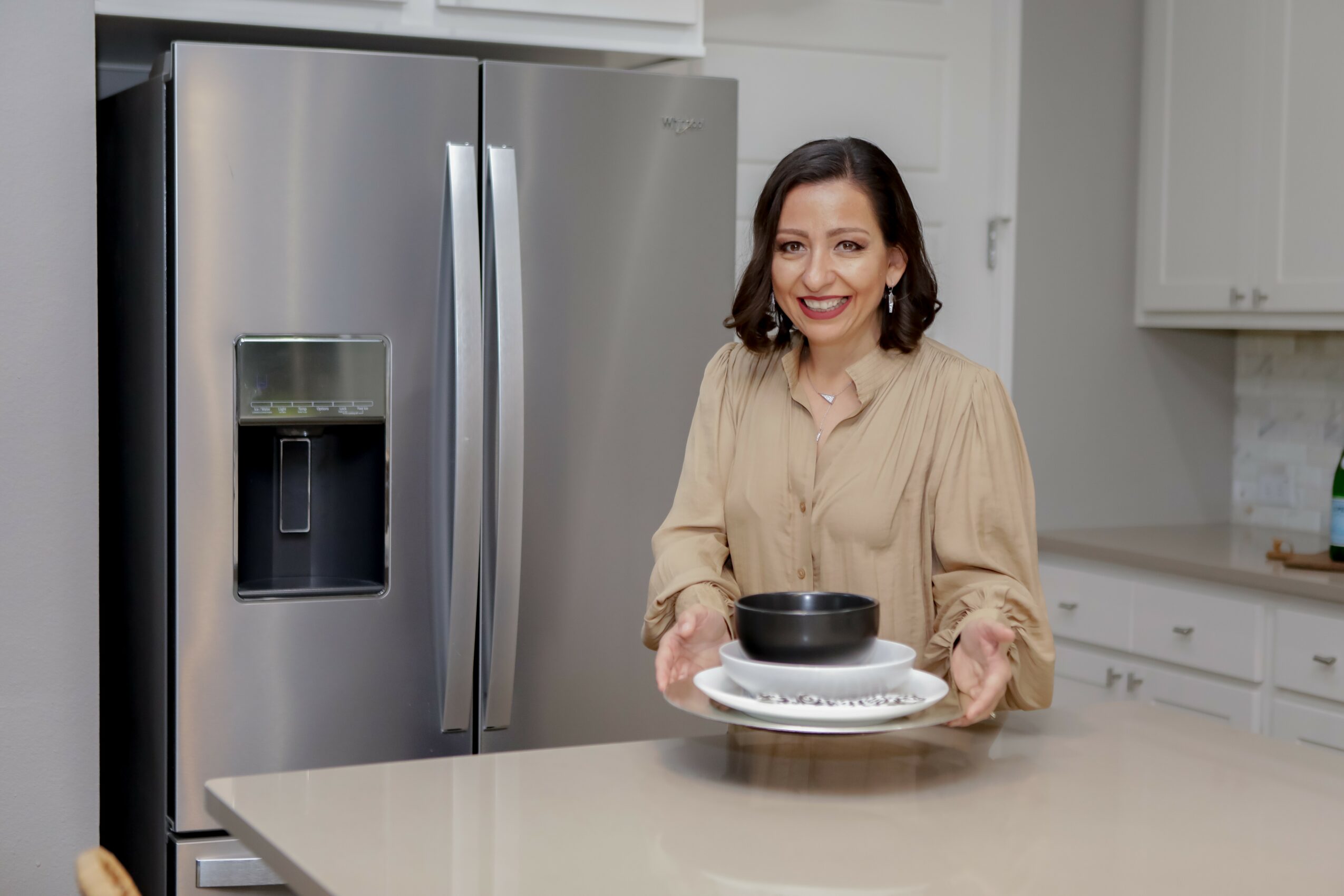 Woman holding a plate with a black bowl in a modern kitchen, smiling, with a stainless steel refrigerator nearby.