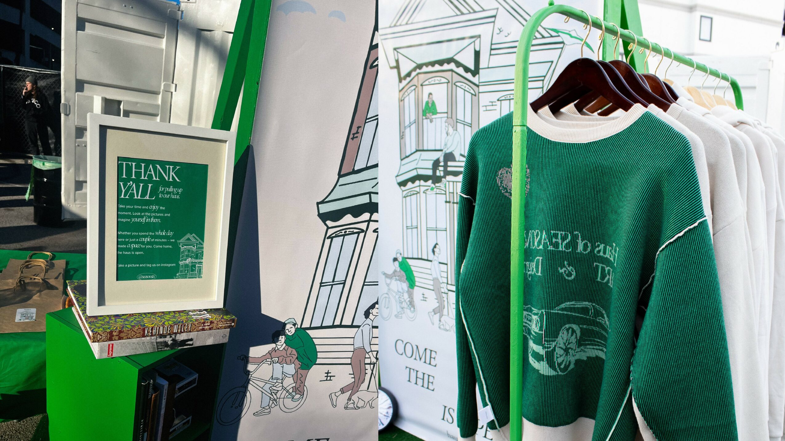 Green sweater hanging on a rack with other clothing, a framed thank you sign, and a drawing of a cityscape in the background.