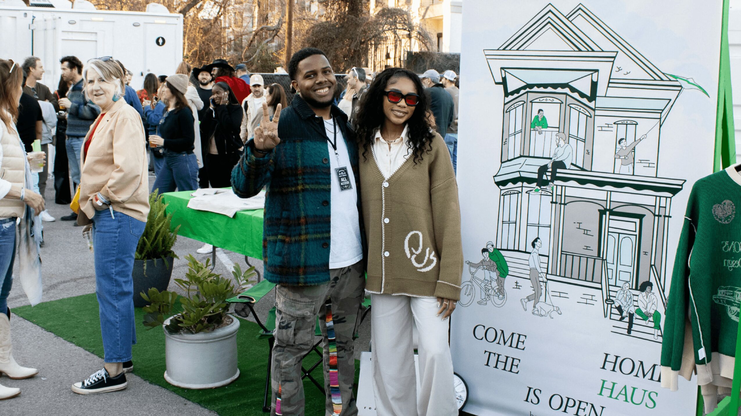 Two people smiling and waving at an outdoor event, with a crowd and a large architectural drawing in the background.
