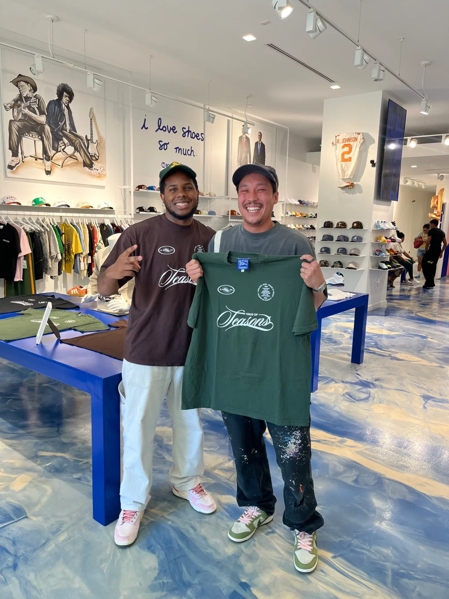 Two smiling men holding a green T-shirt inside a clothing store, with shelves and displays in the background.