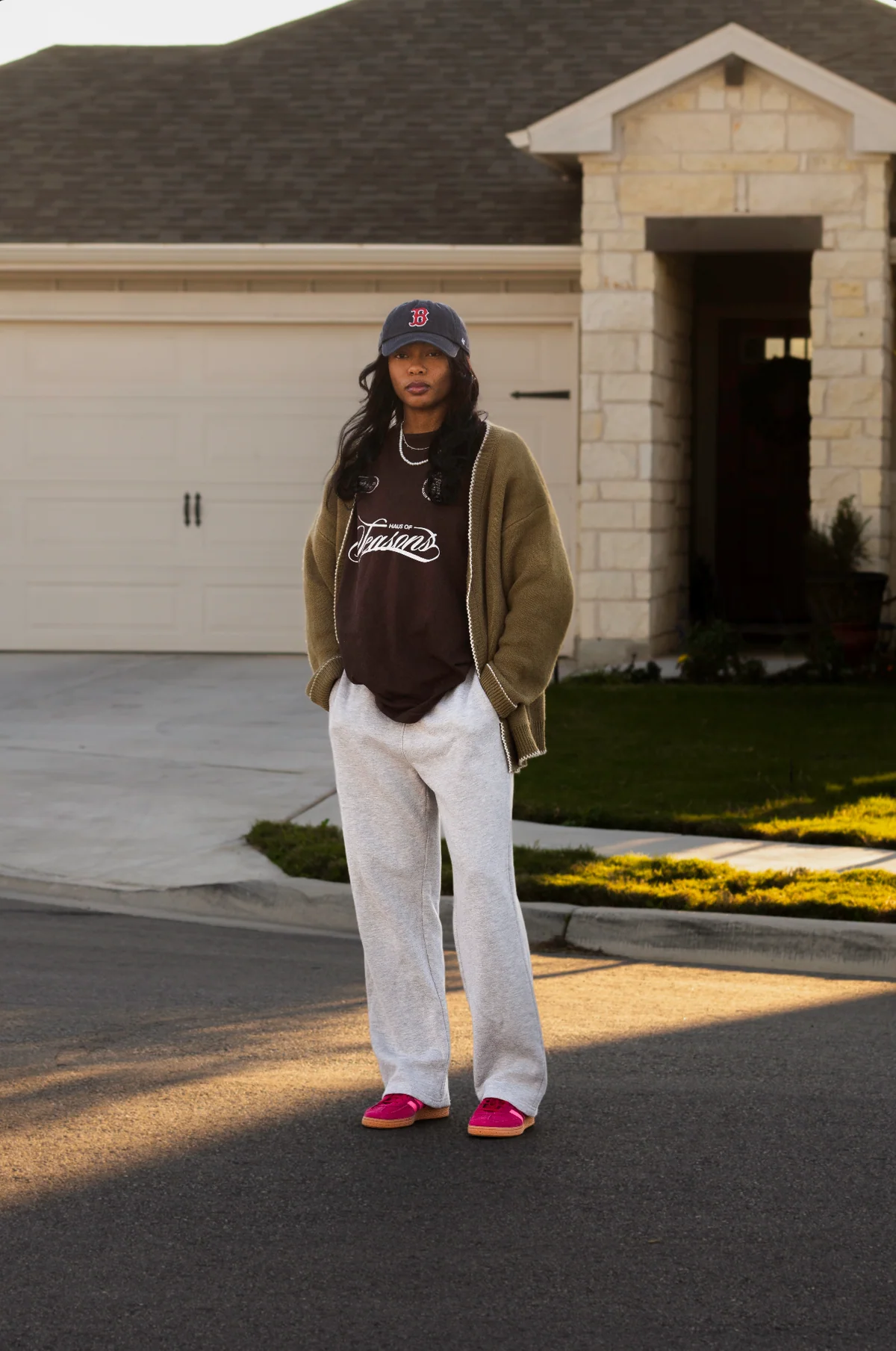 Person standing on street in front of house, wearing a cap, hoodie, and light-colored pants, with pink shoes.