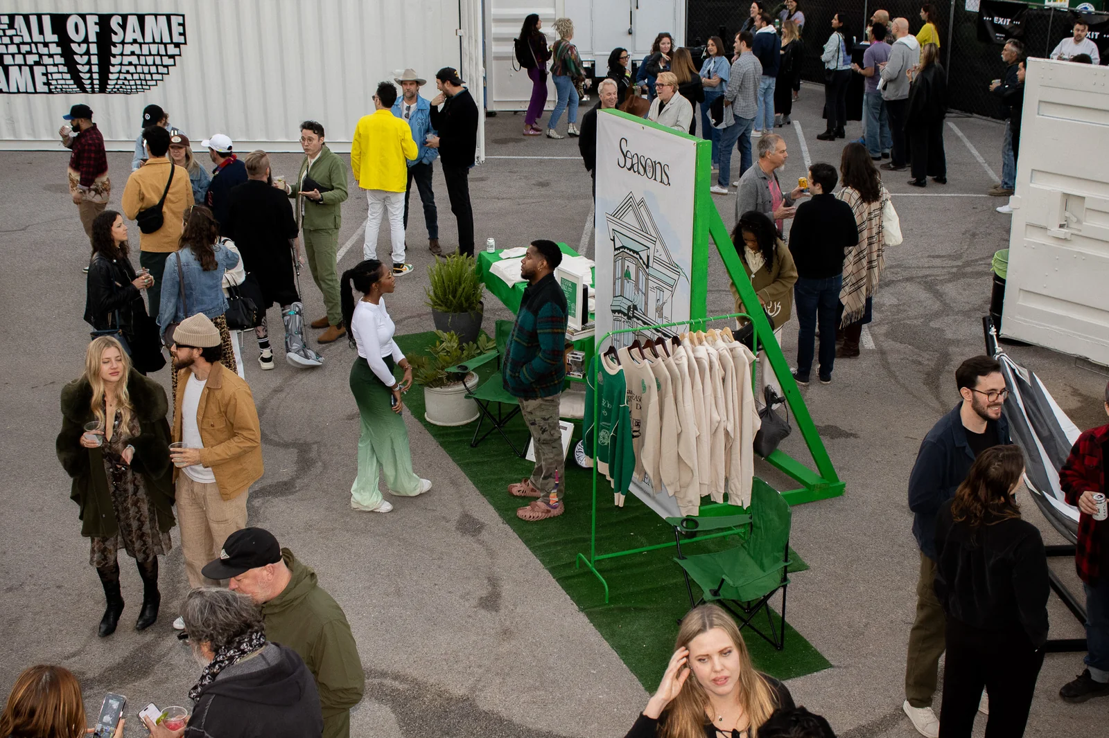 Crowd of people at an indoor event with a green display stand and clothing rack, some walking and talking.