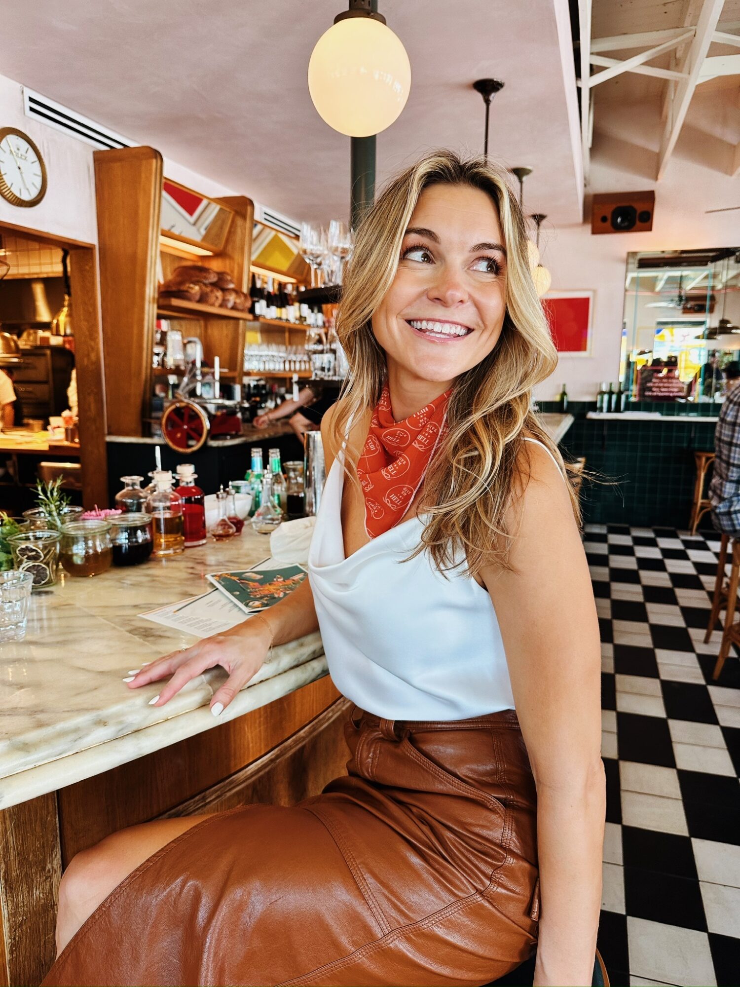 Smiling woman with long hair sitting at a bar counter in a cafe, with shelves of bottles and glasses behind her.
