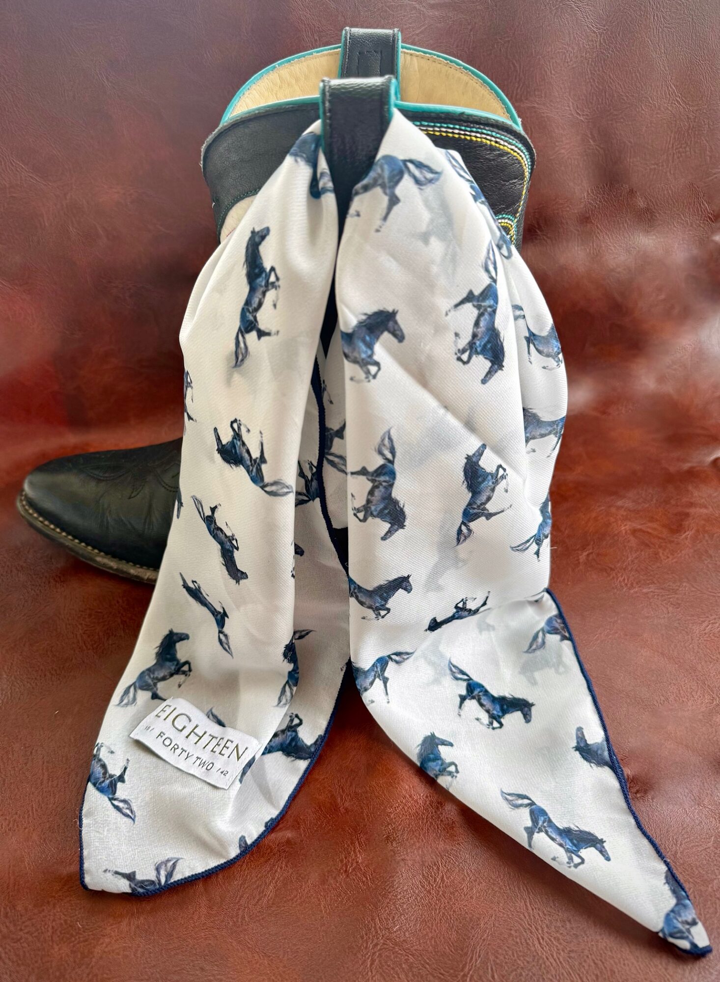 A cowboy hat with a white scarf featuring blue horse patterns draped over it, on a wooden surface.