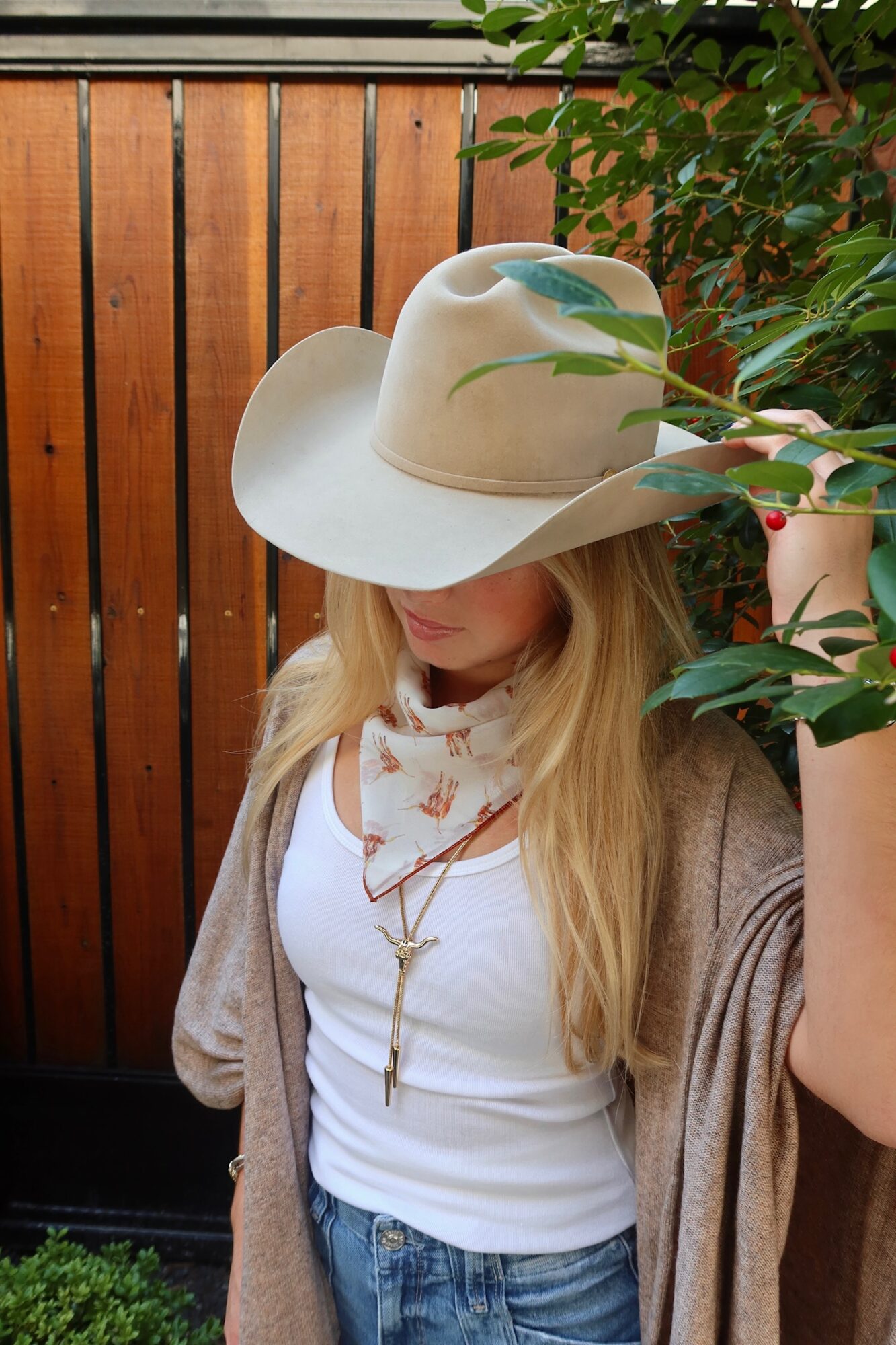 Woman wearing a large hat, white shirt, and beige cardigan, standing near a wooden fence and green plants.