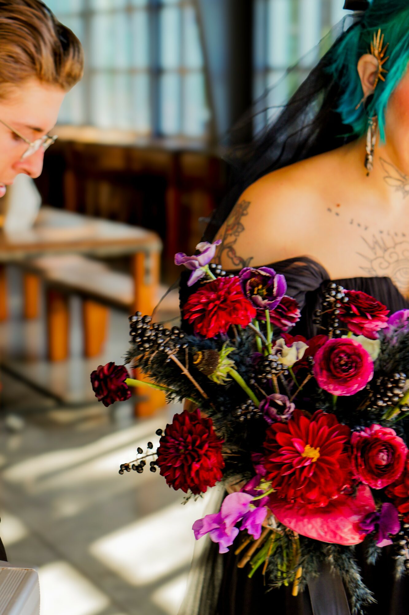 Person with teal hair and black dress holding a large bouquet of red, pink, and purple flowers, indoors with sunlight and wooden furniture.