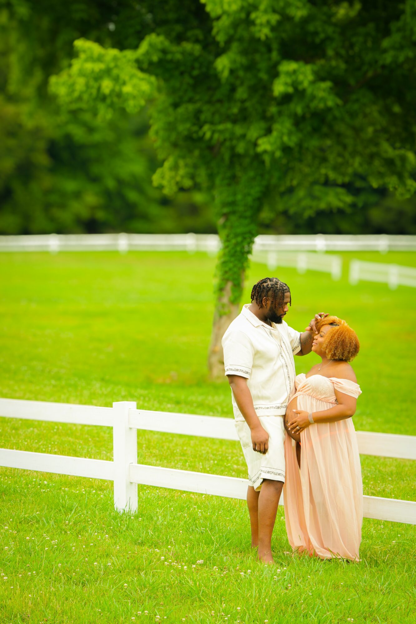 A man and woman stand close together on a grassy field with trees and a white fence in the background.