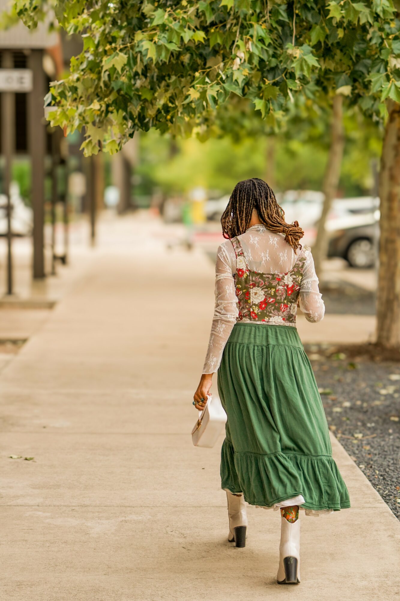 Woman walking on sidewalk with trees and parked cars in background, wearing a floral top and green skirt, holding a bag
