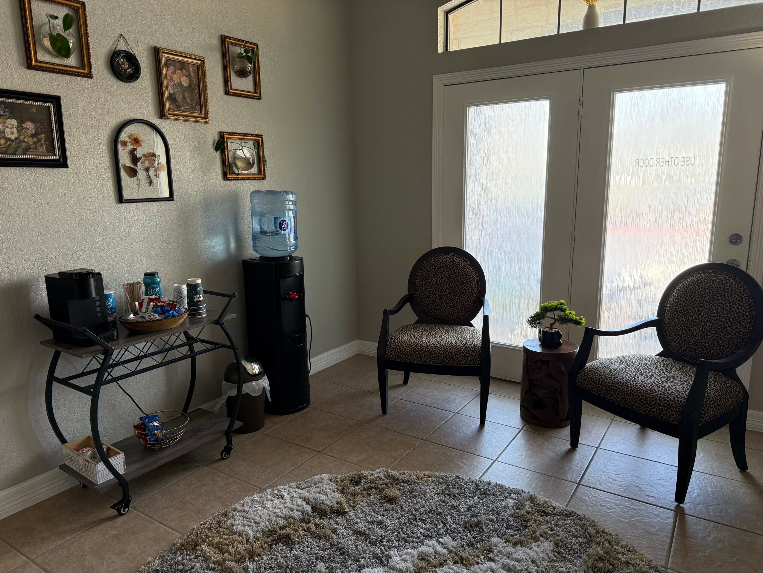 Room with two chairs, a small table with a plant, a water dispenser, and framed pictures on the wall.
