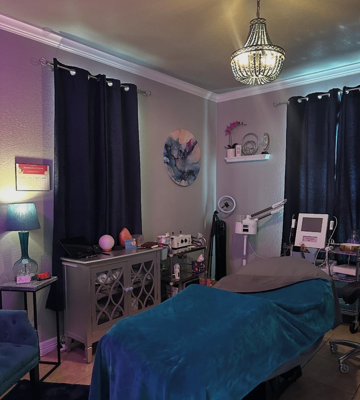 A room with a massage table covered in a blue sheet, dark curtains, a chandelier, and various furniture and equipment.