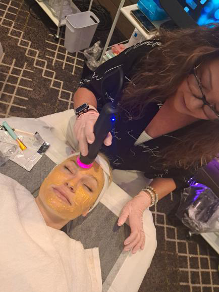 Person receiving a facial treatment with a mask, lying on a bed, while a professional applies a device to their face.