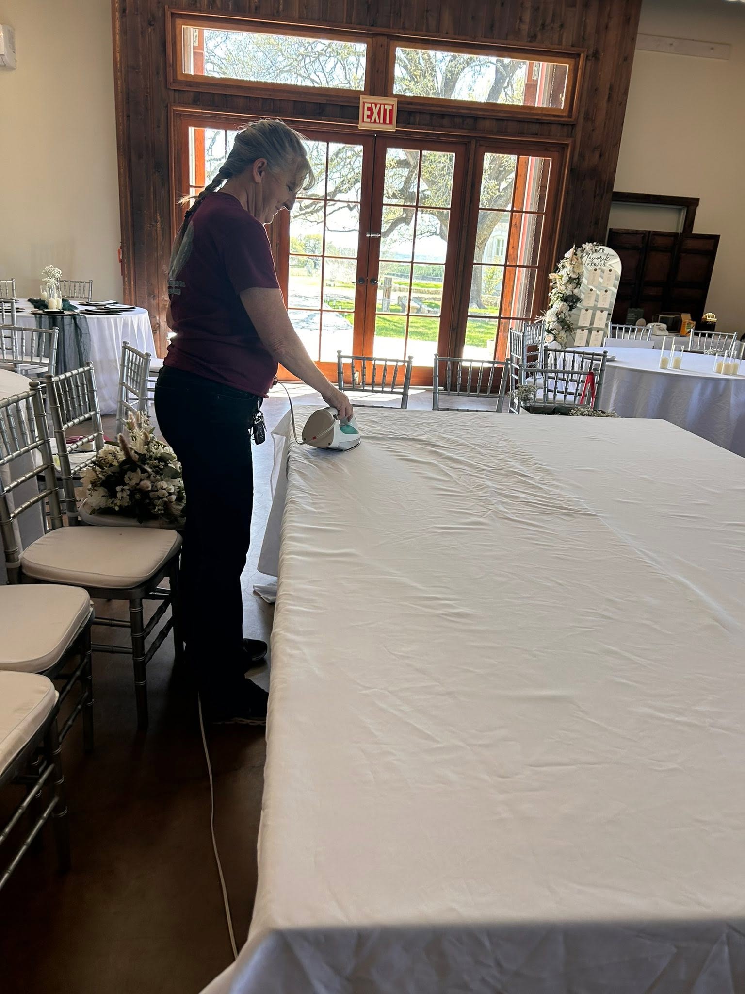 Person standing at a long table, preparing or setting up a white tablecloth in a decorated indoor space with large windows.