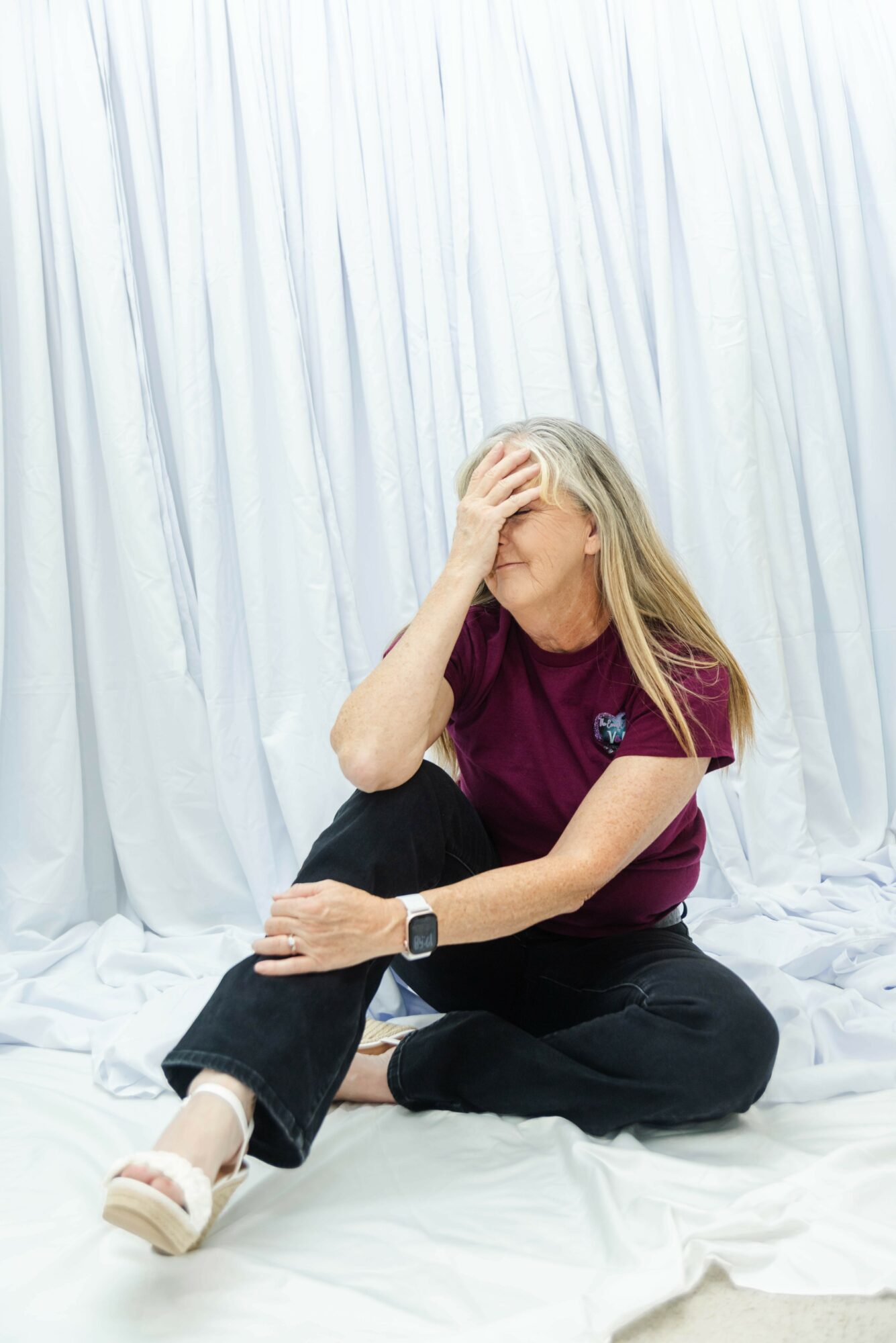 Woman sitting on floor with hand on forehead, white curtain background, wearing maroon shirt and black pants.