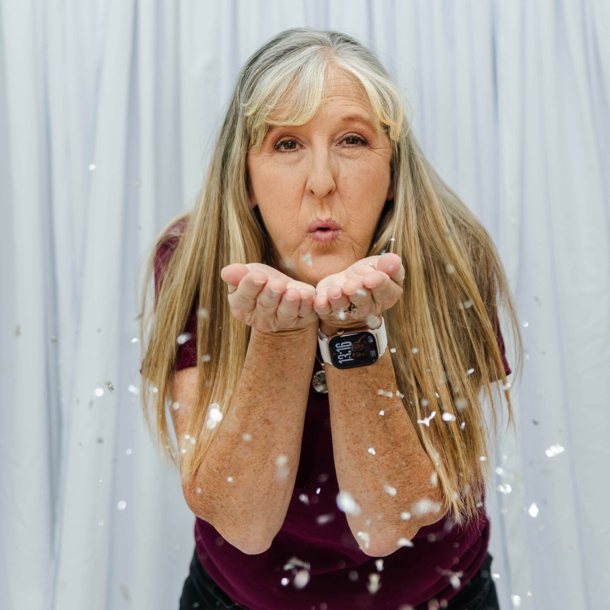 Older woman blowing soap bubbles with a surprised expression, standing in front of a white curtain, wearing a maroon shirt.