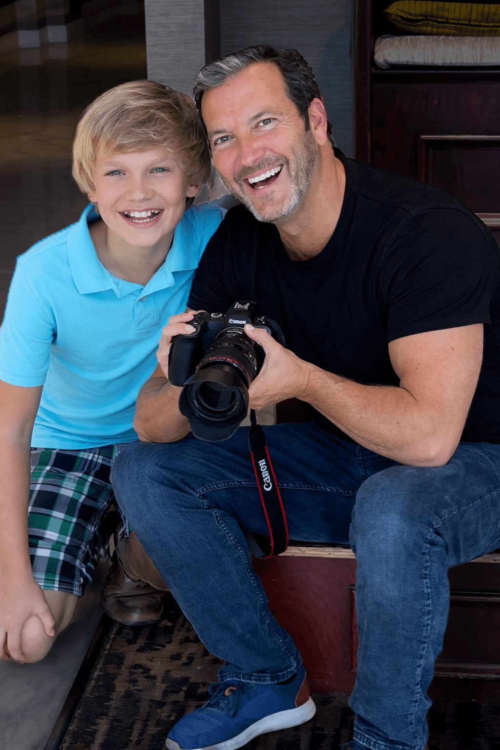 Man and boy smiling, sitting together, man holding a camera, indoors, happy moment, casual clothing