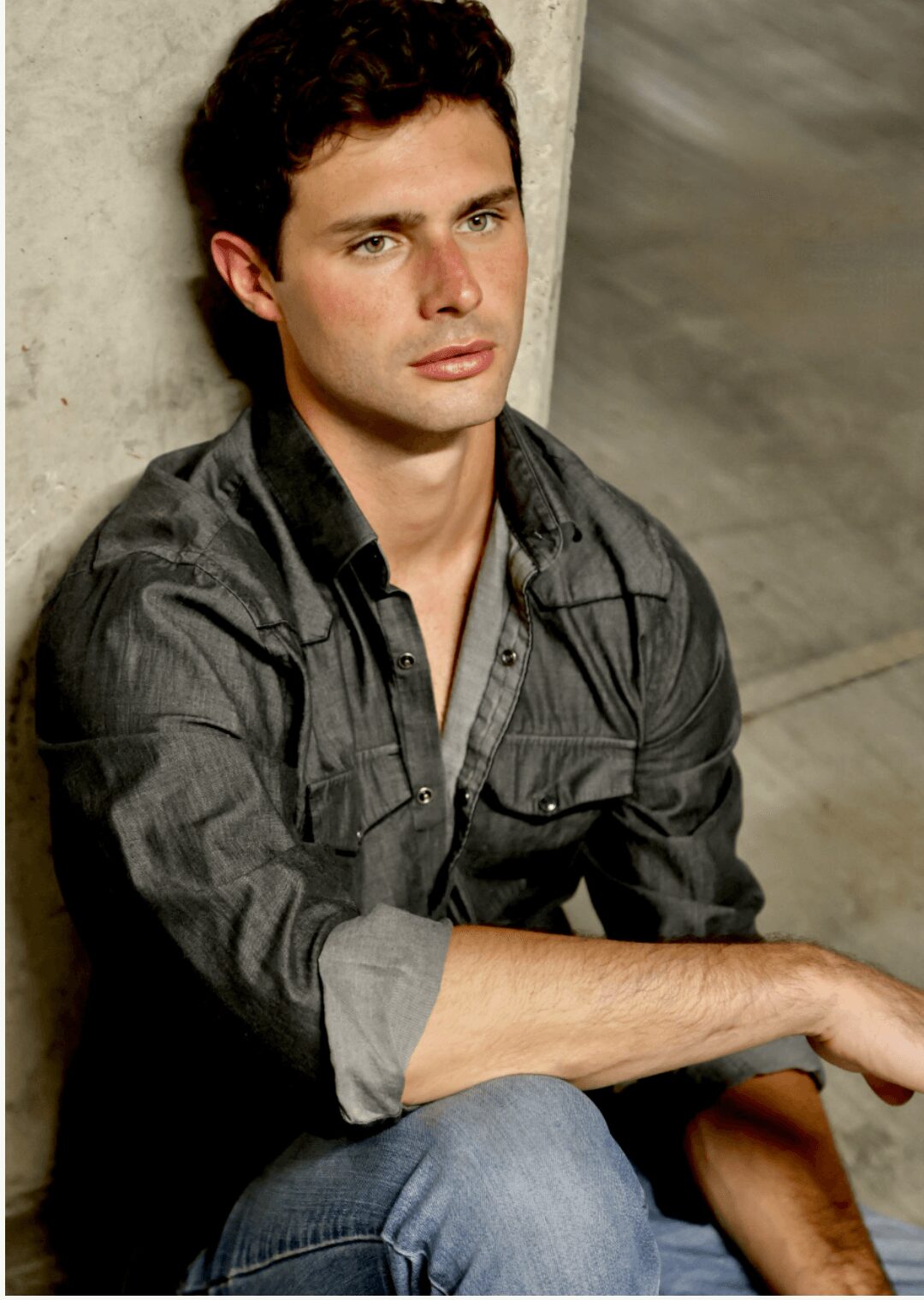 Young man with dark curly hair, wearing a gray shirt, sitting against a concrete wall, looking to the side.