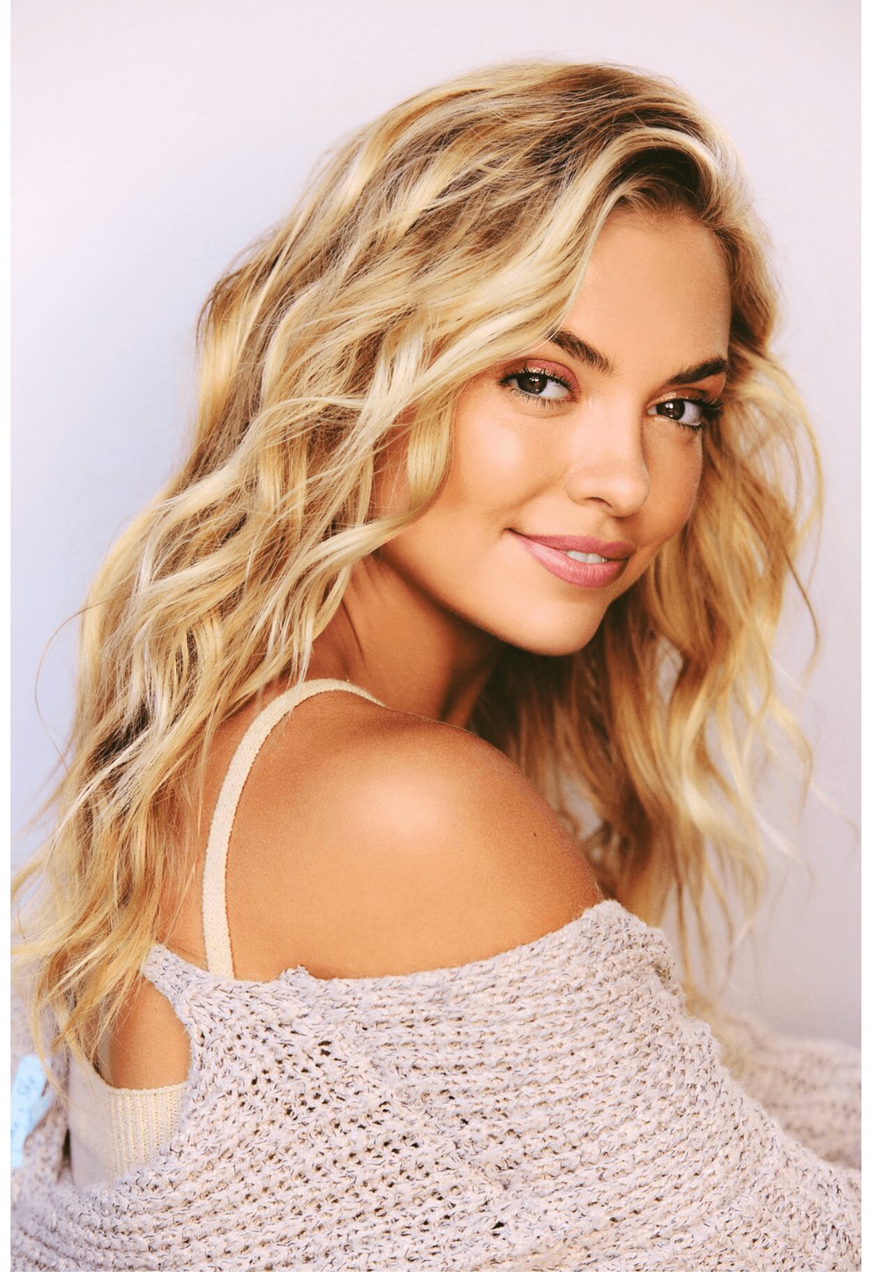 Young woman with wavy blonde hair smiling, wearing a light-colored off-shoulder top, against a plain background.