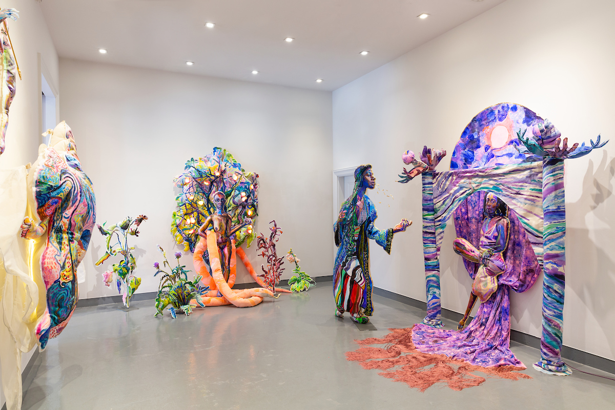Colorful sculptures of people and abstract shapes in an art gallery with white walls and gray floor.