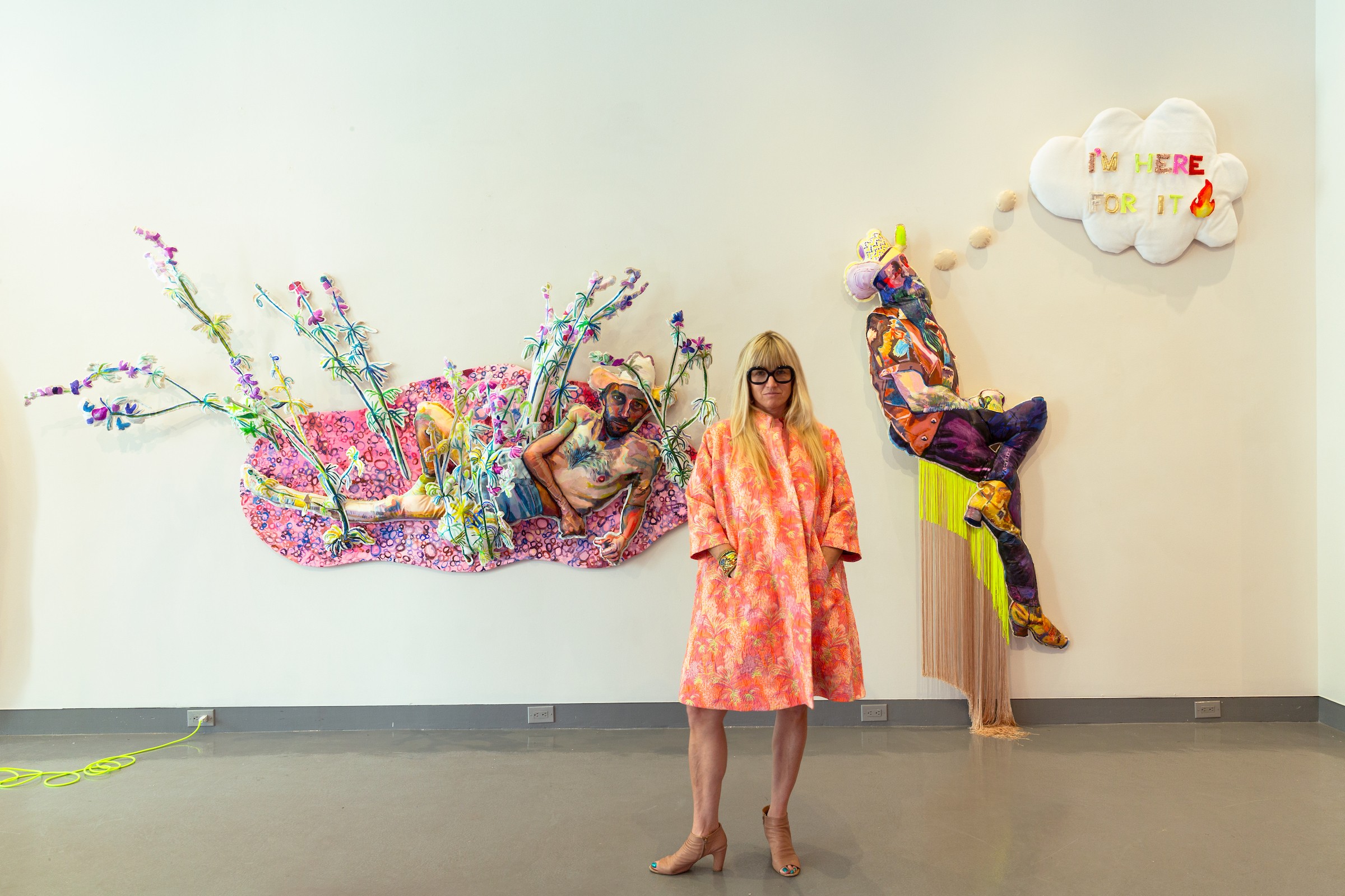 Person standing in front of colorful art installation with two figures and a cloud-shaped sign, plain wall background.