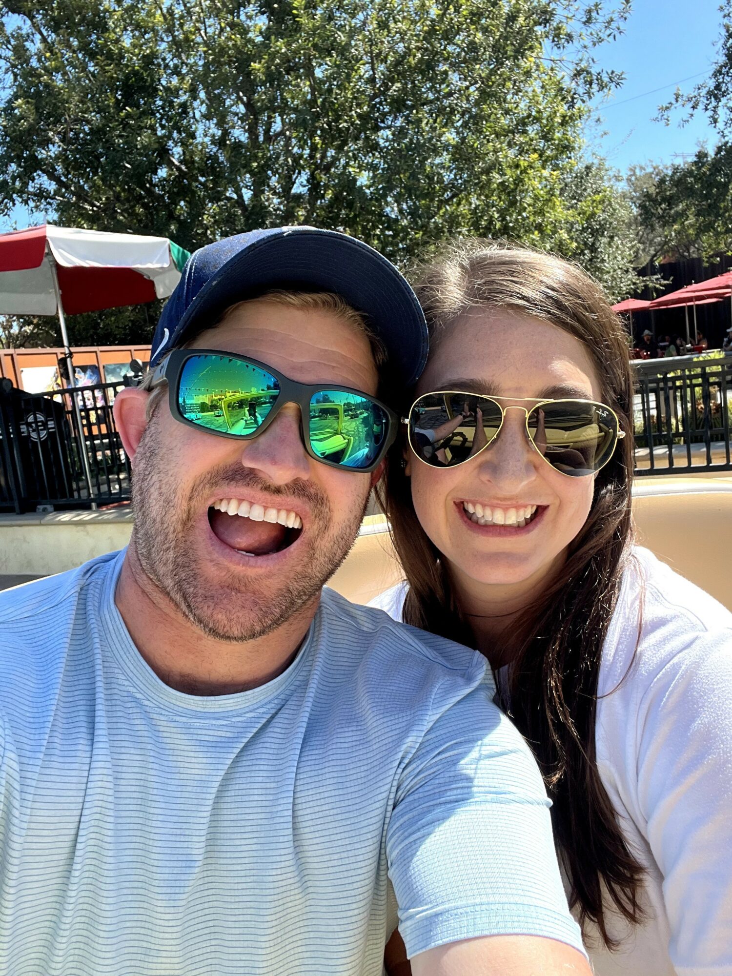 Two people smiling outdoors, wearing sunglasses, with trees and a fence in the background.