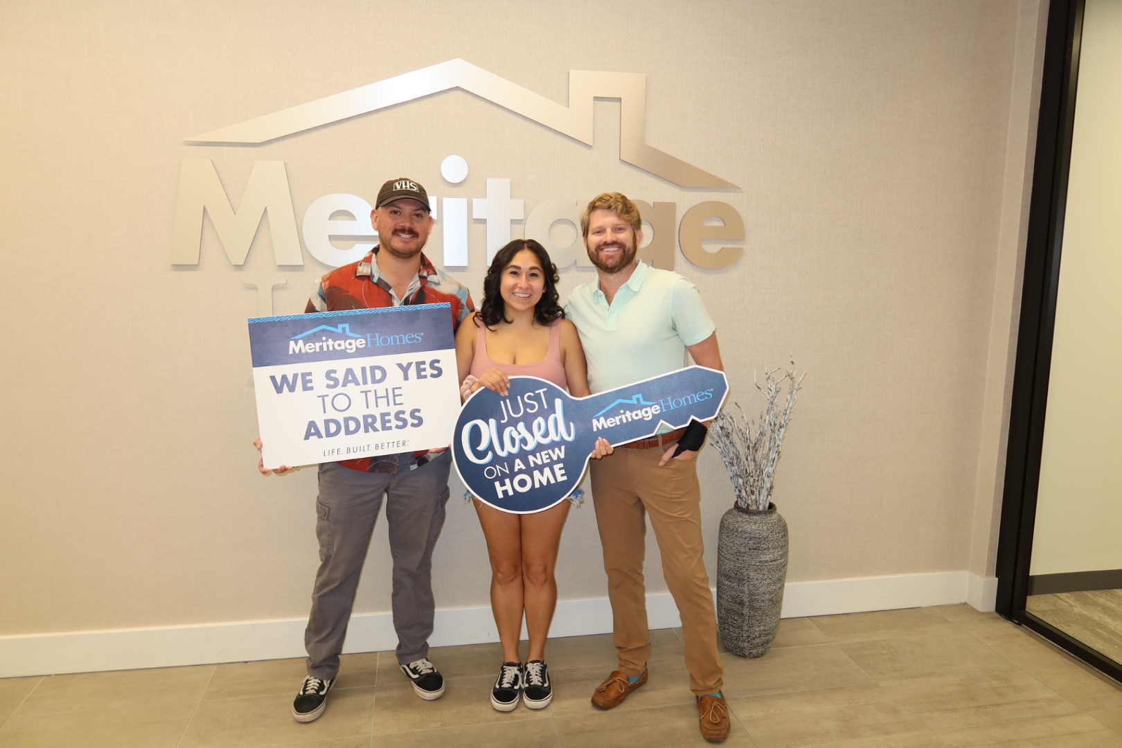 Three people standing indoors holding a large key and a sign, smiling, with a Meritage logo on the wall behind them.